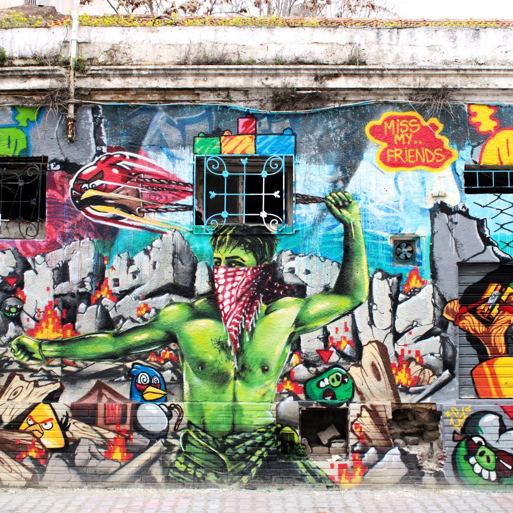 L'immagine mostra un murales colorato su un muro esterno. Al centro, c'è un personaggio verde con un fazzoletto rosso, che sembra esprimere un forte senso di protesta o di resistenza. Sullo sfondo, si possono notare elementi di graffiti e illustrazioni vivaci, tra cui simboli e oggetti in movimento, che creano un'atmosfera dinamica e urbana. Il murales combina diverse tecniche artistiche, rendendolo molto interessante visivamente.