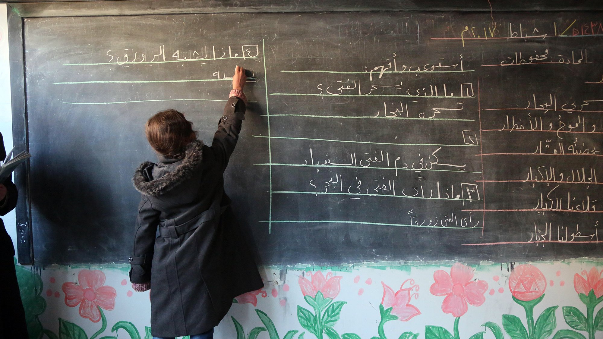 The image depicts a classroom scene where a student is writing on a blackboard. The blackboard has lines of text in Arabic, while the bottom of the board is decorated with colorful flower drawings. The student, wearing a coat, is focused on writing, and there are other individuals in the background, potentially teachers or classmates. The setting appears to be an educational environment.