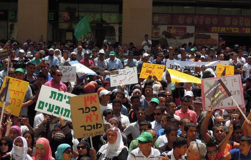 L'image montre une grande foule rassemblée lors d'une manifestation. Les participants tiennent divers panneaux et affiches, certains portant des messages en plusieurs langues, y compris l'hébreu et l'arabe. L'atmosphère semble dynamique, avec des gens exprimant leurs opinions et leurs préoccupations. On peut voir des drapeaux et des bannières, indiquant un fort engagement communautaire et un appel à l'action.