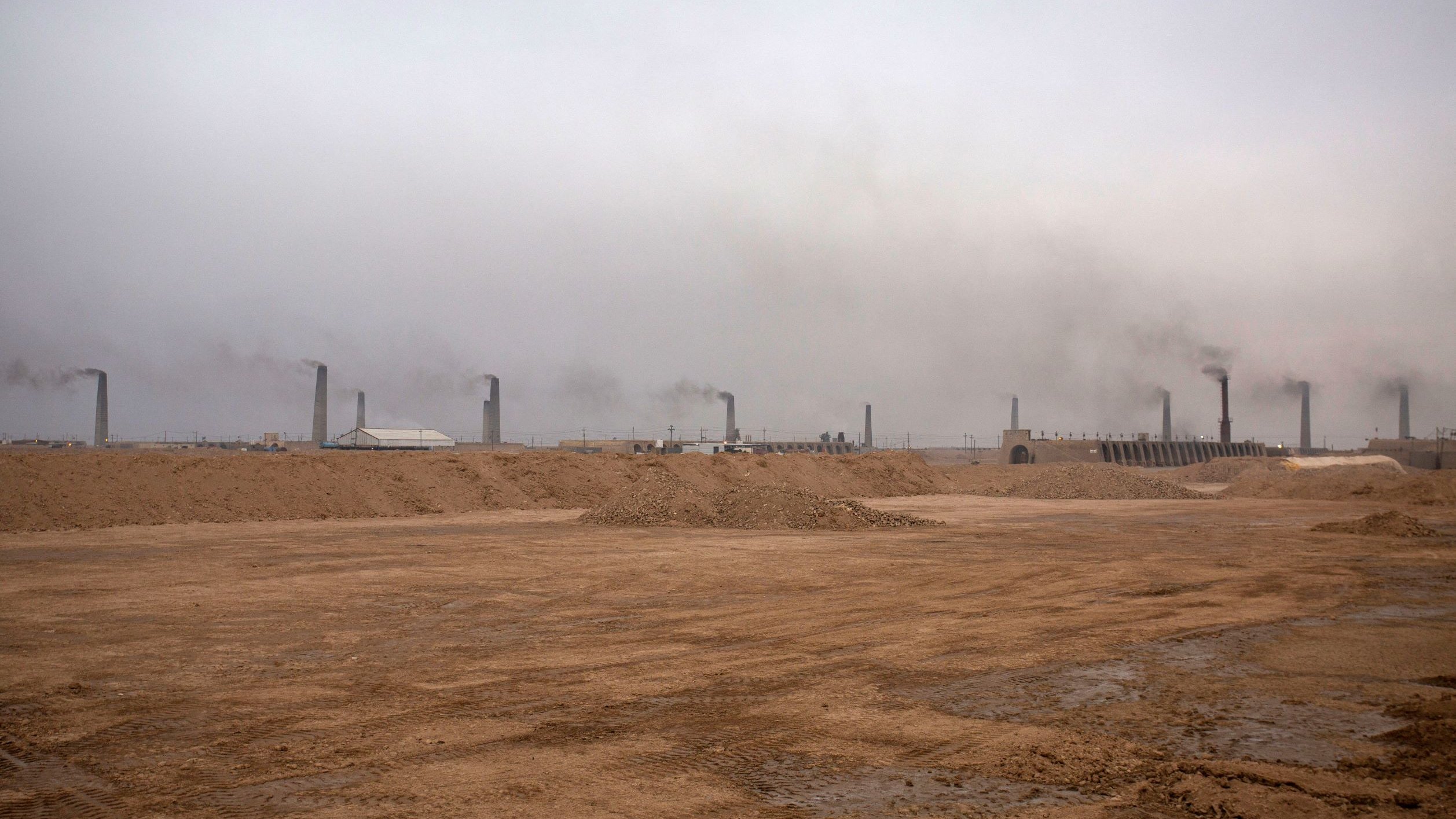 تظهر الصورة منظرًا لموقع صناعي في منطقة صحراوية، حيث يهيمن على المشهد ضباب كثيف أو سحب من الدخان تتصاعد من مداخن المنشآت. الأرض تظهر جافة وغير مستوية، مع كميات من الرمل والتربة. الأجواء تبدو رمادية وموحشة، مما يعكس تأثير النشاط الصناعي على البيئة.
