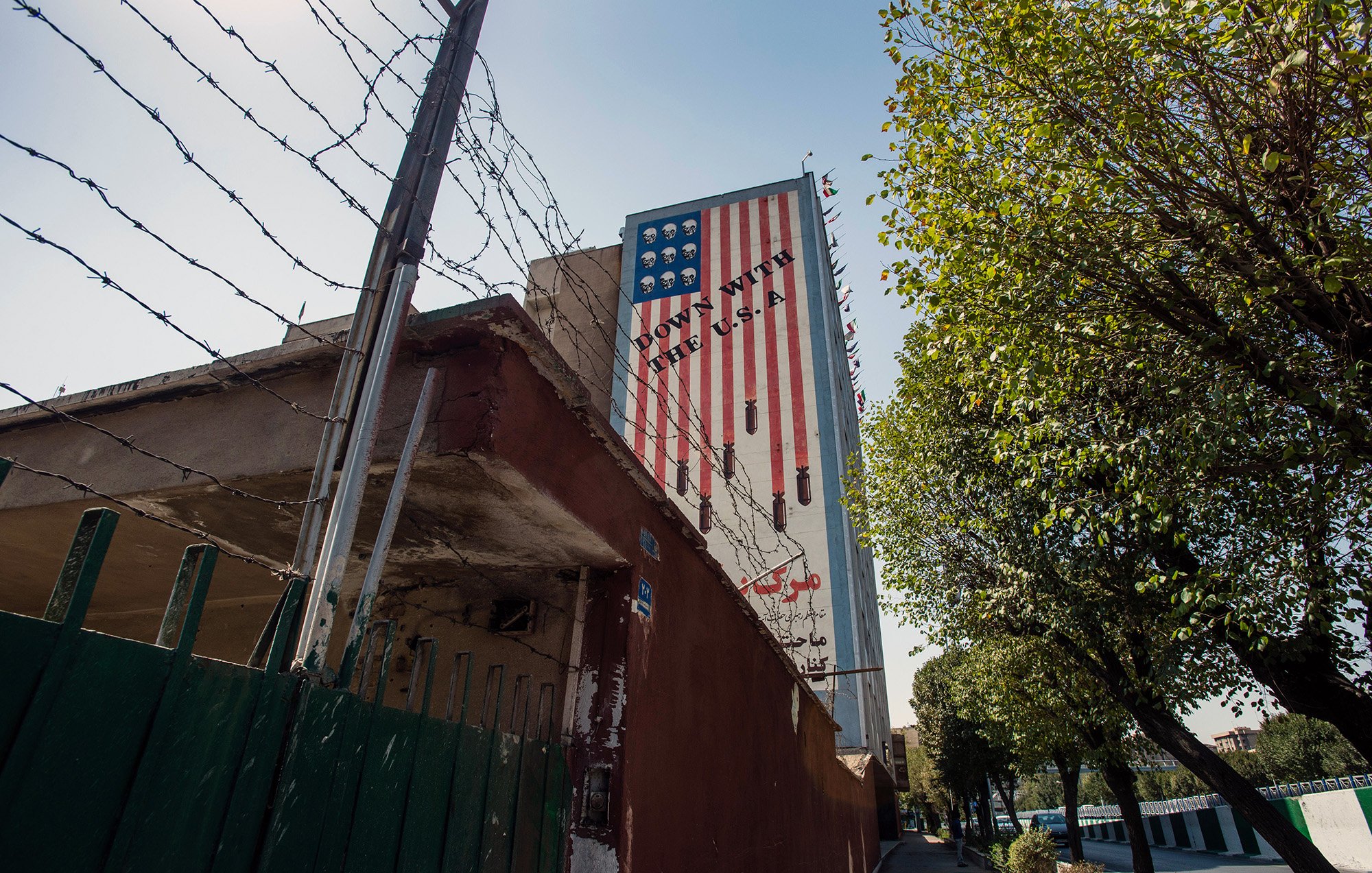 تصویر نشان‌دهنده دیواری است که بر روی آن پرچم آمریکا نقاشی شده است. این پرچم به گونه‌ای طراحی شده که بخشی از آن به صورت معکوس و غیرمتعارف نمایش داده شده است. در پس‌زمینه، fences (حصارهایی) با میله‌های تیز دیده می‌شود. همچنین درختان و یک پیاده‌رو در کنار دیوار وجود دارد. نور آفتاب به تصویر زیبایی افزوده است و فضا به طور کلی حس خاصی از تاریخ و سیاست را منتقل می‌کند.