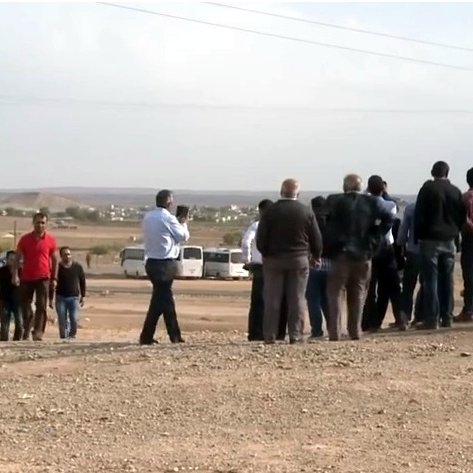 L'image montre un groupe de personnes rassemblées sur un terrain semi-aride. Ils semblent observer quelque chose au loin, peut-être un événement ou une scène particulière. L'arrière-plan est dégagé, avec des collines et des bâtiments visibles au loin. Il y a des véhicules stationnés, dont une voiture à droite. L'atmosphère suggère une interaction entre les gens, qui semblent concentrés sur ce qu'ils regardent. Des câbles électriques sont visibles, ajoutant à l'ambiance rurale de la scène.