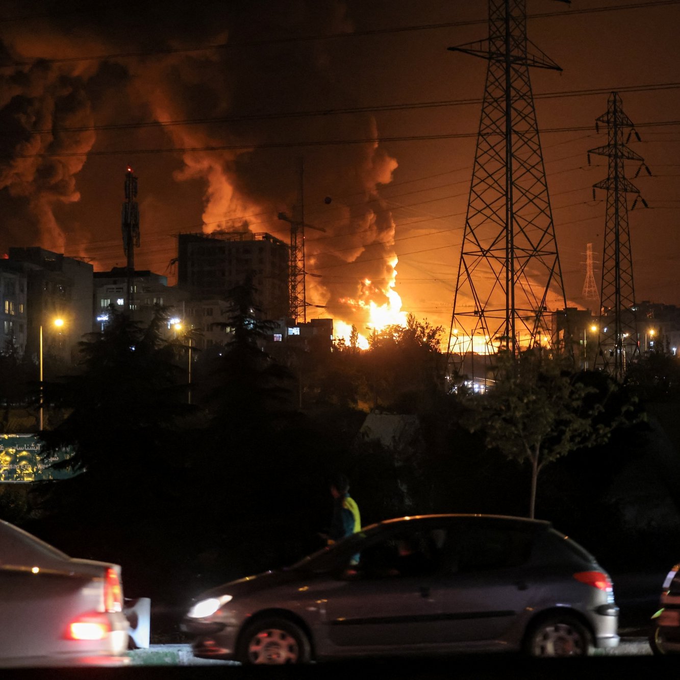 Bright flames and dark smoke rise from buildings at night, with cars passing nearby.