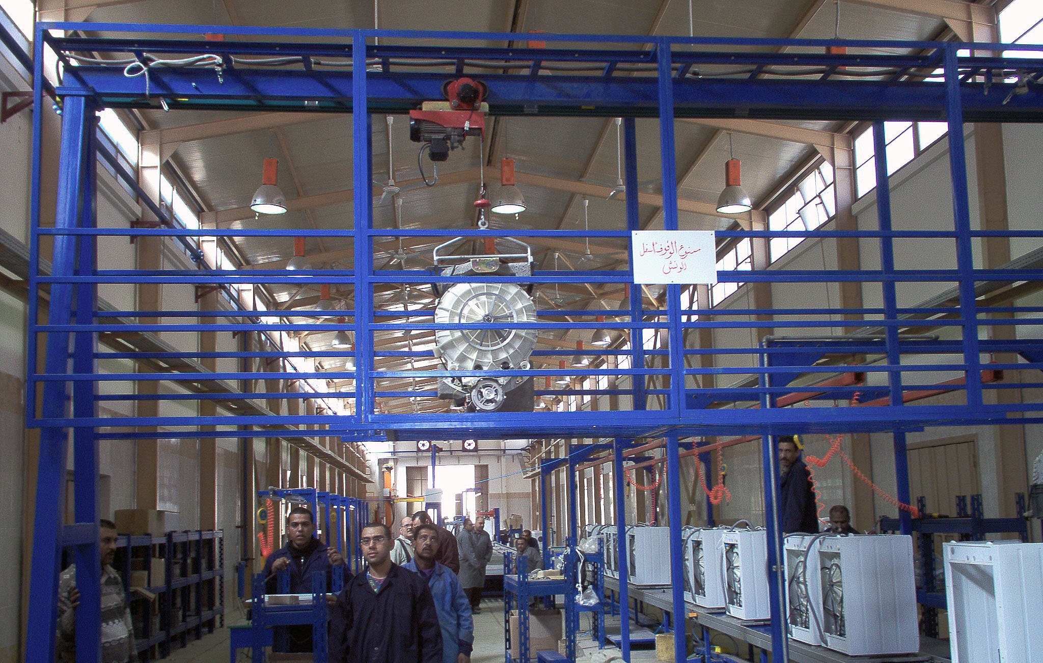 الصورة تبين مشهداً داخل مصنع أو ورشة عمل. يظهر في الصورة عدة أشخاص يعملون في بيئة صناعية. توجد آلات وأجهزة كهربائية مرتبة على طاولات أو أسطح عمل، بالإضافة إلى نظام تعليقي يحمل بعض المعدات في السقف. الإضاءة جيدة، مما يساهم في خلق جو عمل مناسب. كما يوجد لافتة معلقة على الجدار تحمل نصاً، مما يشير إلى وجود توجيهات أو معلومات مهمة في المكان.