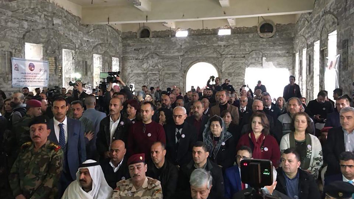 L'image montre une grande foule de personnes rassemblées dans un bâtiment à l'apparence industrielle ou historique, avec des murs en pierre. Les participants semblent être assis ou debout et portent des badges ou des pin's. Diverses personnes de différentes origines apparaissent, y compris des représentants militaires en uniforme. L'atmosphère semble formelle, suggérant un événement significatif ou une réunion. Des caméras et des journalistes sont également présents, indiquant qu'il pourrait s'agir d'une conférence ou d'une célébration.
