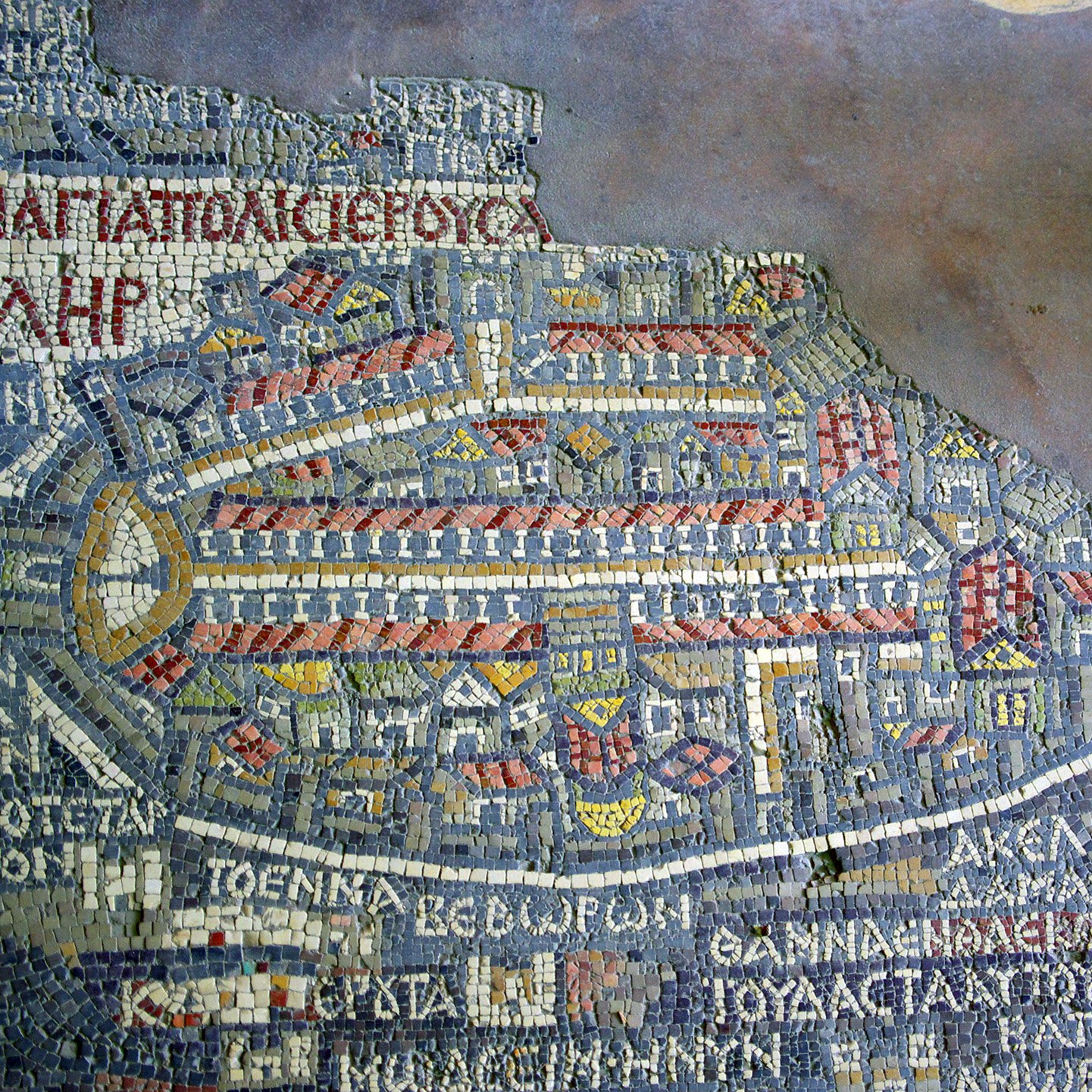 The image depicts a mosaic map, featuring intricate tile work that illustrates an ancient city layout. The map is composed of various colored tiles, forming buildings, streets, and outlines of the architecture within the city. It includes labeled regions or areas, indicating places of significance in the city, which might be historical or prominent during the time it represents. The overall design reflects a detailed representation of urban planning, showcasing the artistry and craftsmanship of the era.