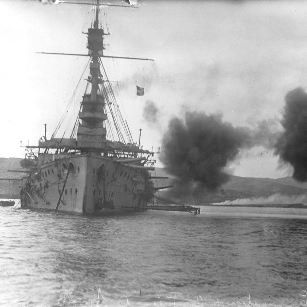 L'image montre un grand navire de guerre, probablement un cuirassé, tirant des armes à feu, ce qui crée des nuages de fumée sur l'eau. Le navire est équipé de plusieurs canons sur ses flancs. En arrière-plan, on aperçoit des collines ou des montagnes, ainsi qu'une éventuelle base maritime ou un autre type d'installations côtières. L'atmosphère est dynamique, accentuée par l'action de tir.
