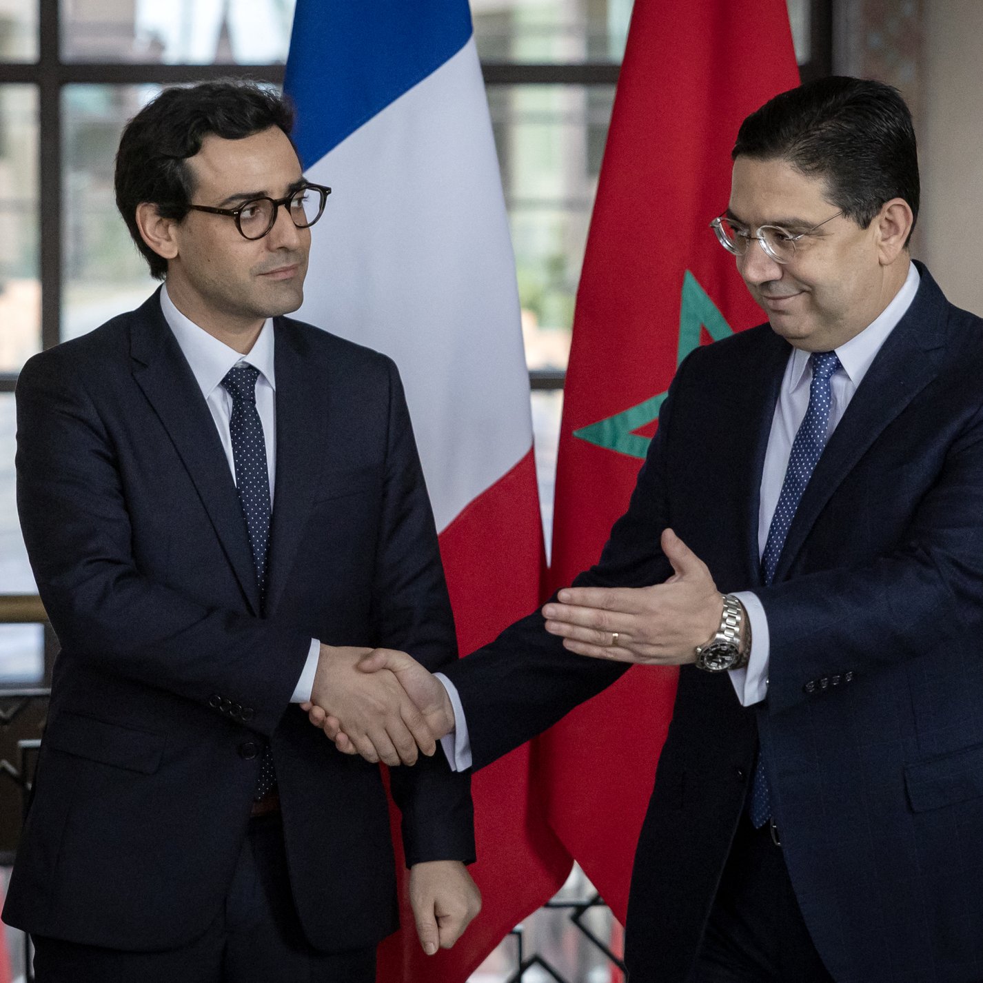 تظهر الصورة رجلين يرتديان بدلات رسمية. أحدهما يقف على اليمين ويمثل المغرب، حيث يظهر علم المغرب خلفه. الآخر يقف على اليسار ويمثل فرنسا، مع العلم الفرنسي خلفه. يبدو أن الرجلين يتصافحان في سياق رسمي، مما يشير إلى اجتماع أو تعاون بين البلدين. القاعة التي يتواجدان بها تبدو أنيقة وتحتوي على نوافذ كبيرة تسمح بدخول الضوء الطبيعي.