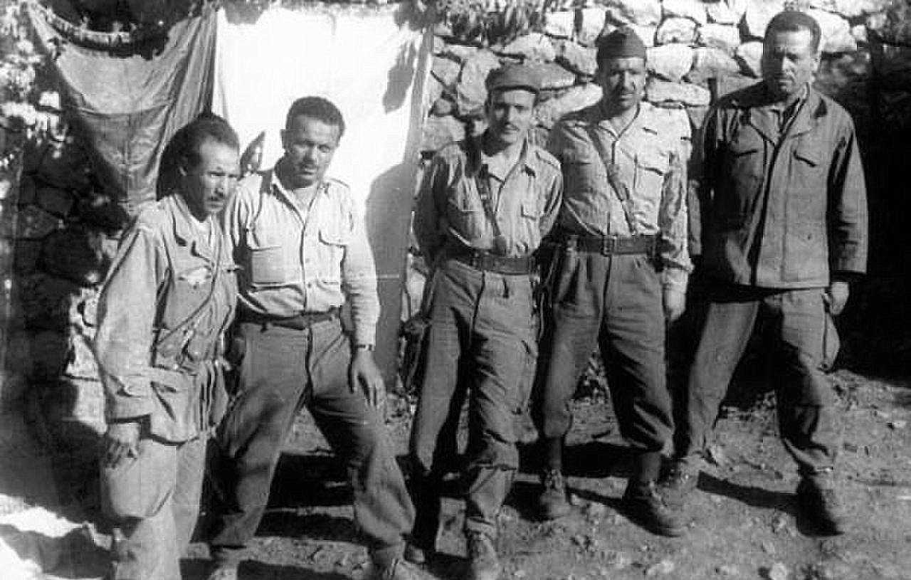 The image shows a group of five soldiers standing together in a rural setting. They are wearing military uniforms and appear to be posing for the photo. The background consists of a stone wall and a makeshift tarp or cloth, suggesting a temporary encampment. The soldiers have serious expressions, reflecting the context of their environment. Overall, the image conveys a sense of camaraderie among them in a military setting.