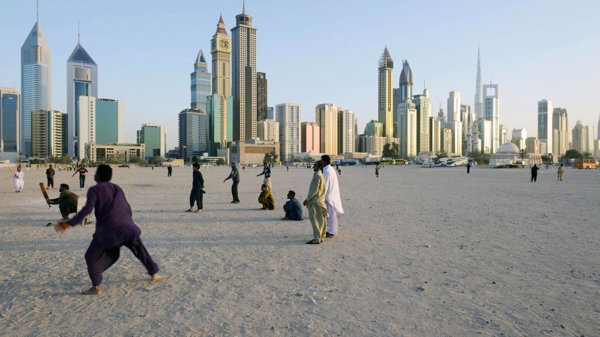 L'image montre une plage ou un espace ouvert, avec des gens qui se déplacent ou jouent dans le sable. En arrière-plan, on peut voir une ville moderne avec de grands gratte-ciel, représentant une skyline impressionnante. Les bâtiments semblent contemporains et variés en termes d'architecture. L'éclairage indique qu'il s'agit probablement d'une fin de journée, avec un ciel clair. L'atmosphère semble animée, avec des personnes profitant du lieu.