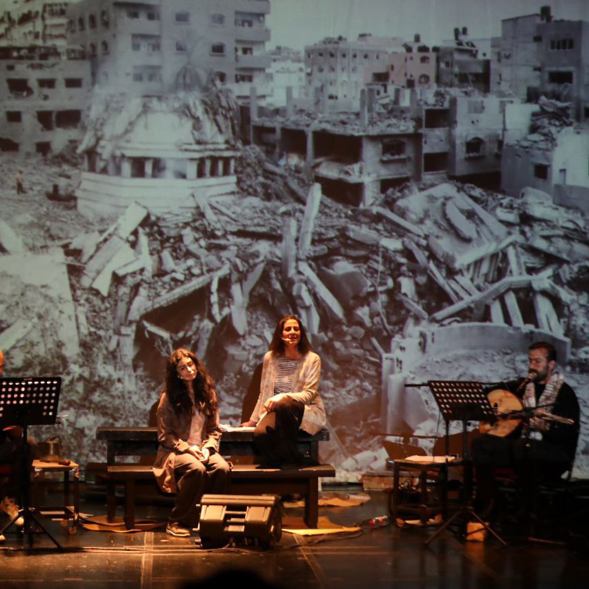 Four performers sit with instruments in front of a large backdrop of war-torn buildings.