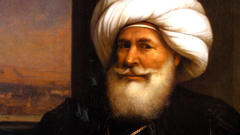L'image représente un homme portant une tenue traditionnelle, avec un turban distinctif et une barbe blanche. Il a un sourire visible et semble confiant. L'homme est assis, tenant une épée décorative dans sa main, ce qui suggère une certaine dignité ou autorité. En arrière-plan, on peut apercevoir un paysage, probablement représentant une scène architecturale ou naturelle. L'ensemble donne une impression de grandeur et de culture.