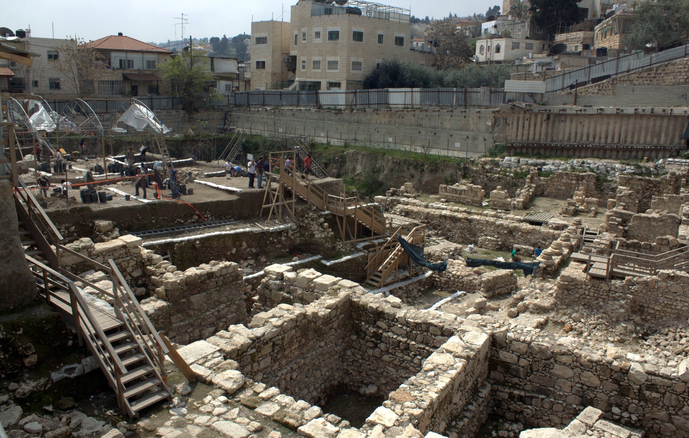 L'image montre des fouilles archéologiques en cours dans une zone urbaine. On peut voir des structures en pierre disposées de manière organisée, avec des vestiges de murs et de fondations visibles. Des scaffolds et des passerelles en bois sont installés pour permettre aux archéologues de travailler et d'accéder facilement aux différentes couches de sol. En arrière-plan, des bâtiments modernes sont présents, ce qui témoigne du mélange entre le patrimoine historique et l'urbanisation contemporaine. Des chercheurs et des travailleurs s'activent autour du site, suggérant une recherche active sur l'histoire de la région.