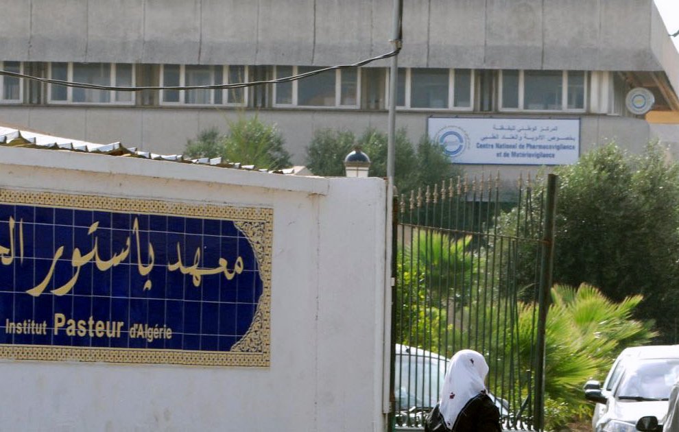 L'image montre l'extérieur de l'Institut Pasteur en Algérie. On peut voir un bâtiment moderne en arrière-plan avec des panneaux d'affichage. À l'avant-plan, il y a une entrée avec une façade blanche où figure le nom de l'institut en caractères arabes et français. Quelques personnes marchent dans cette zone, suggérant un environnement fréquenté. Des arbres et de la végétation ajoutent une touche de verdure au décor.