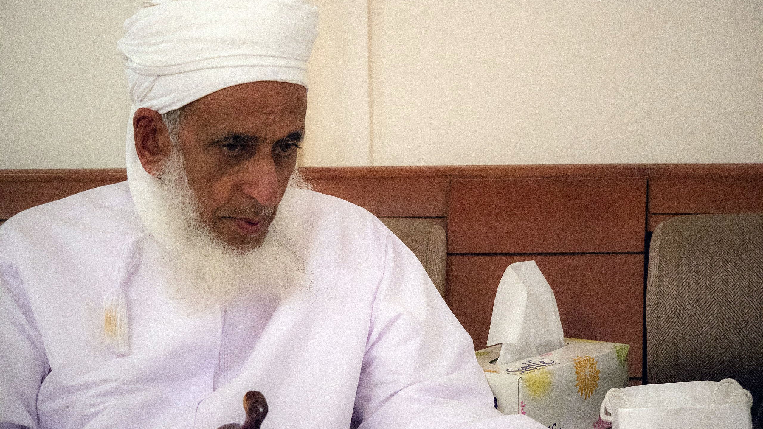 L'image montre un homme âgé portant un turban blanc et une robe traditionnelle. Il a une barbe grise et semble pensif, assis sur un canapé. À côté de lui, on peut voir des mouchoirs et un sac en papier, ce qui suggère qu'il se trouve dans un environnement intérieur calme. L'éclairage est doux, créant une atmosphère sereine.