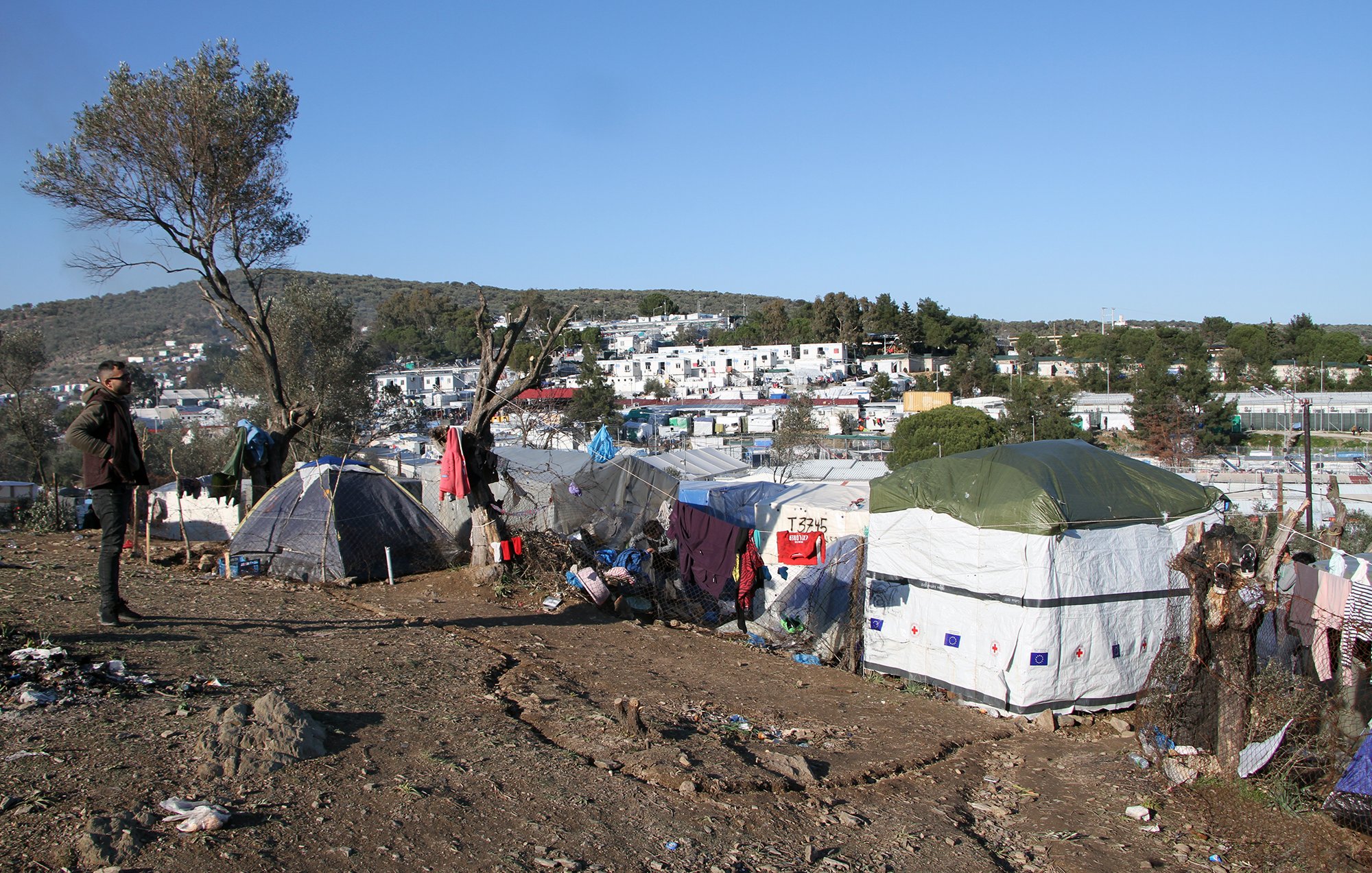 The image depicts a makeshift settlement in a rural or semi-urban area. Numerous tents are arranged on a slope, suggesting a temporary living situation. The landscape features sparse vegetation and a few trees, while in the background, there are more established buildings, possibly residences or facilities. The scene conveys a sense of hardship, with signs of disarray and poverty surrounding the tents. The sky is clear with little cloud cover, indicating a sunny day. A person stands in the foreground, observing the surroundings.