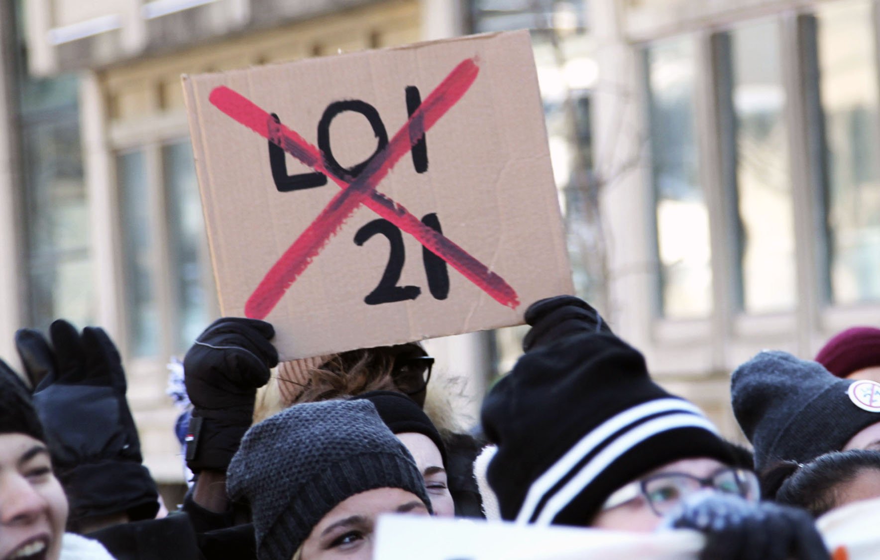 تظهر الصورة مجموعة من الأشخاص في تجمع أو احتجاج، حيث يحمل أحدهم لافتة تشير إلى معارضة لشيء ما. اللافتة مكتوبة عليها "21" مع علامة "إكس" حمراء تدل على رفض هذا الرقم أو القانون. يبدو أن المشاركين يرتدون ملابس شتوية، مما يوحي أن الحدث وقع في فصل الشتاء. تعكس الصورة روح الحماس والمشاركة من قبل المحتجين.
