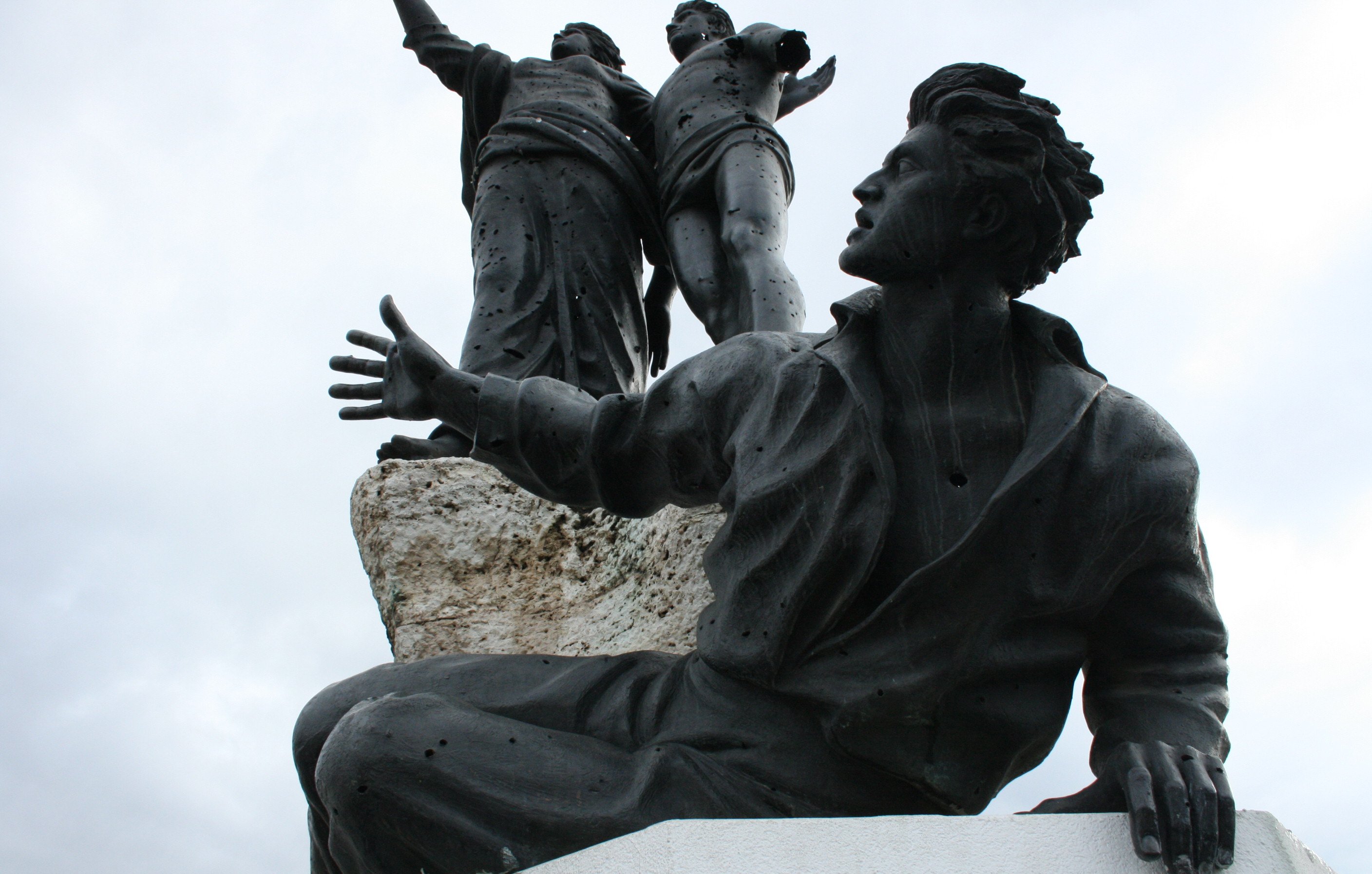 L'image montre une sculpture en bronze représentant plusieurs figures humaines. Au premier plan, un homme est assis, les bras ouverts, avec une expression qui semble transmettre une émotion forte. Derrière lui, deux figures debout sont également visibles, levant les bras, ce qui pourrait suggérer un message de célébration ou de libération. La sculpture est placée sur une base rocheuse, ajoutant une dimension texturisée à l'ensemble. Le ciel apparaît nuageux, ce qui donne un ton dramatique à la scène.