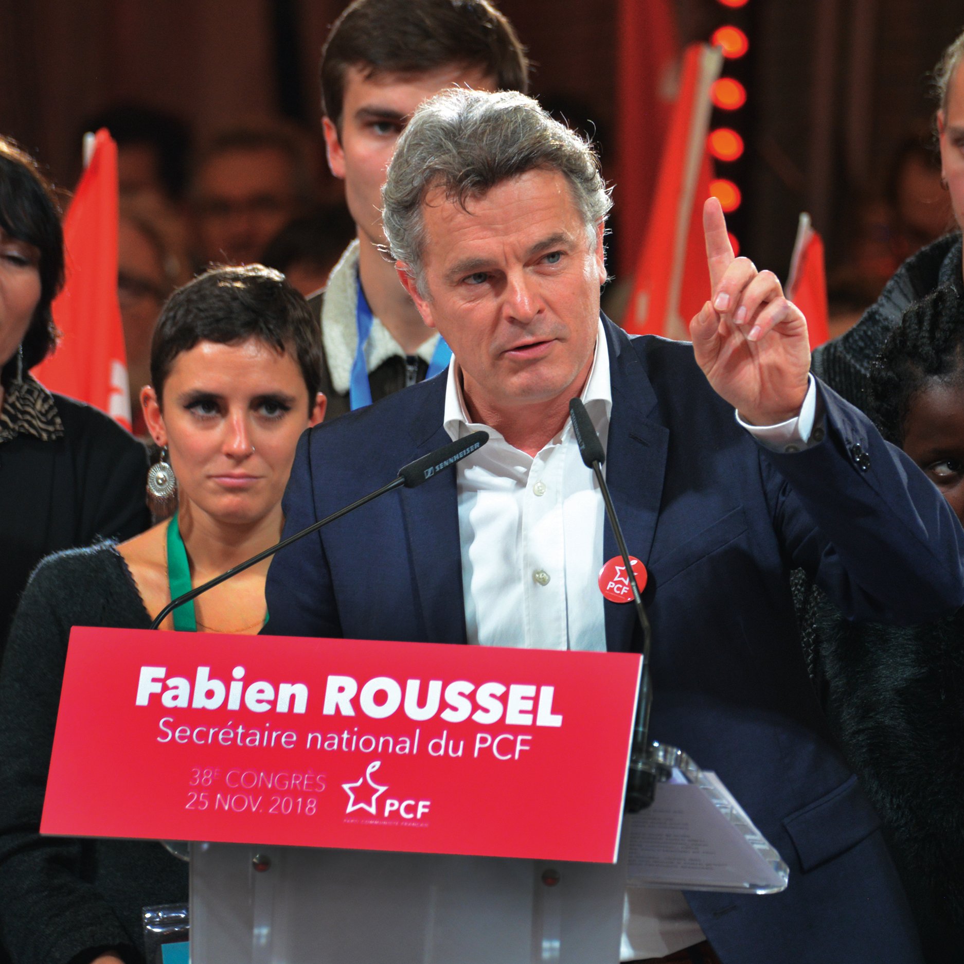 L'image montre un homme au premier plan, probablement en train de faire un discours. Il porte une veste et a l'air déterminé, levant un doigt comme s'il soulignait un point important. Derrière lui, plusieurs personnes l'écoutent, certaines regardant directement l'objectif. En bas de l'image, on peut voir une pancarte rouge avec le texte « Fabien ROUSSEL, Secrétaire national du PCF » et la date du 25 novembre 2018. L'ambiance semble être celle d'un congrès ou d'un rassemblement politique.