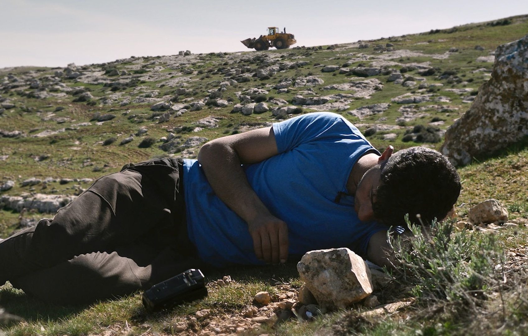 L'image montre un homme allongé sur le sol dans un paysage rural. Il semble être en train de se reposer ou de dormir, la tête posée sur son bras. Ses vêtements sont simples, et il est entouré de roches et de végétation. En arrière-plan, on aperçoit une pelleteuse ou un engin de chantier qui travaille sur le terrain. Le ciel est dégagé, ce qui donne une atmosphère calme à la scène, bien que la présence de l'engin suggère une activité en cours.