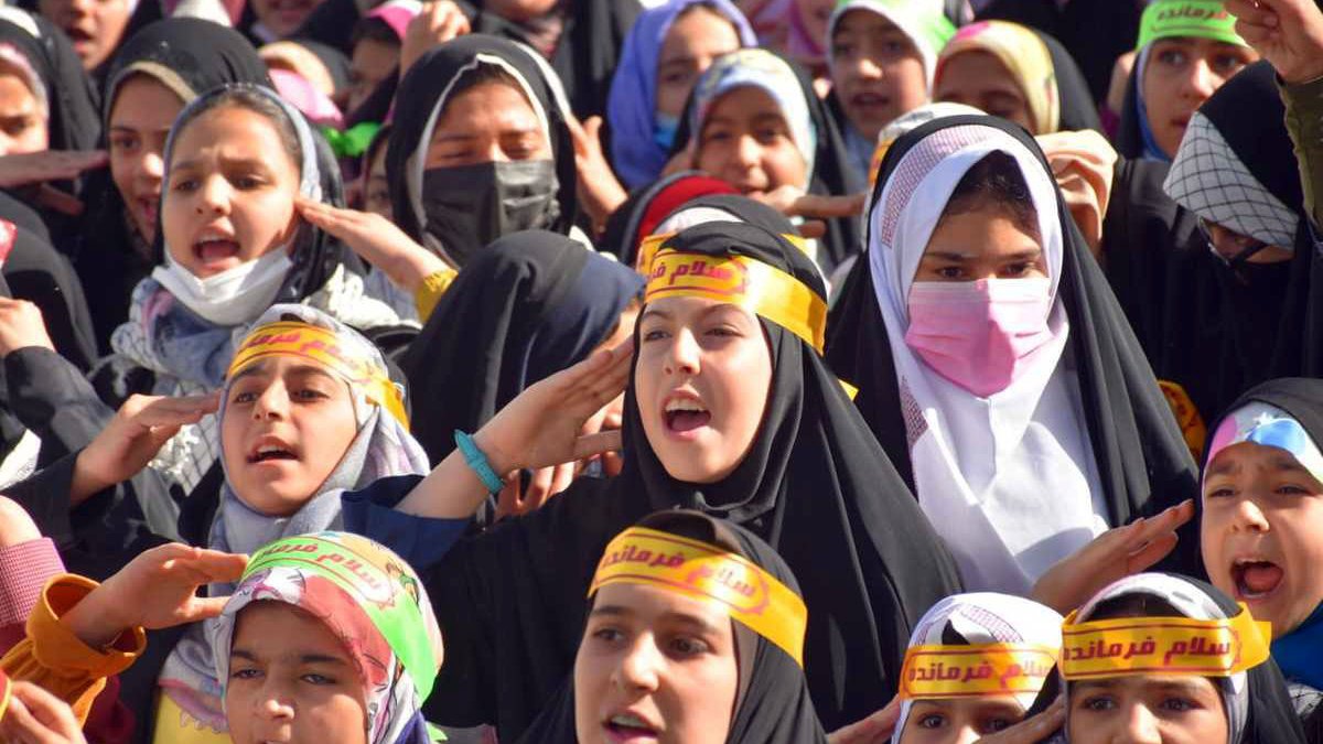 L'image montre un grand groupe de jeunes filles, portant des hijabs et des bandeaux avec des inscriptions en arabe. Elles semblent exprimer de l'enthousiasme, chantant ou criant, et leurs visages sont pleins d'énergie. L'arrière-plan indique un rassemblement, probablement lié à un événement spécial ou une manifestation. Les couleurs des vêtements et des bandeaux apportent une atmosphère vibrante à la scène.