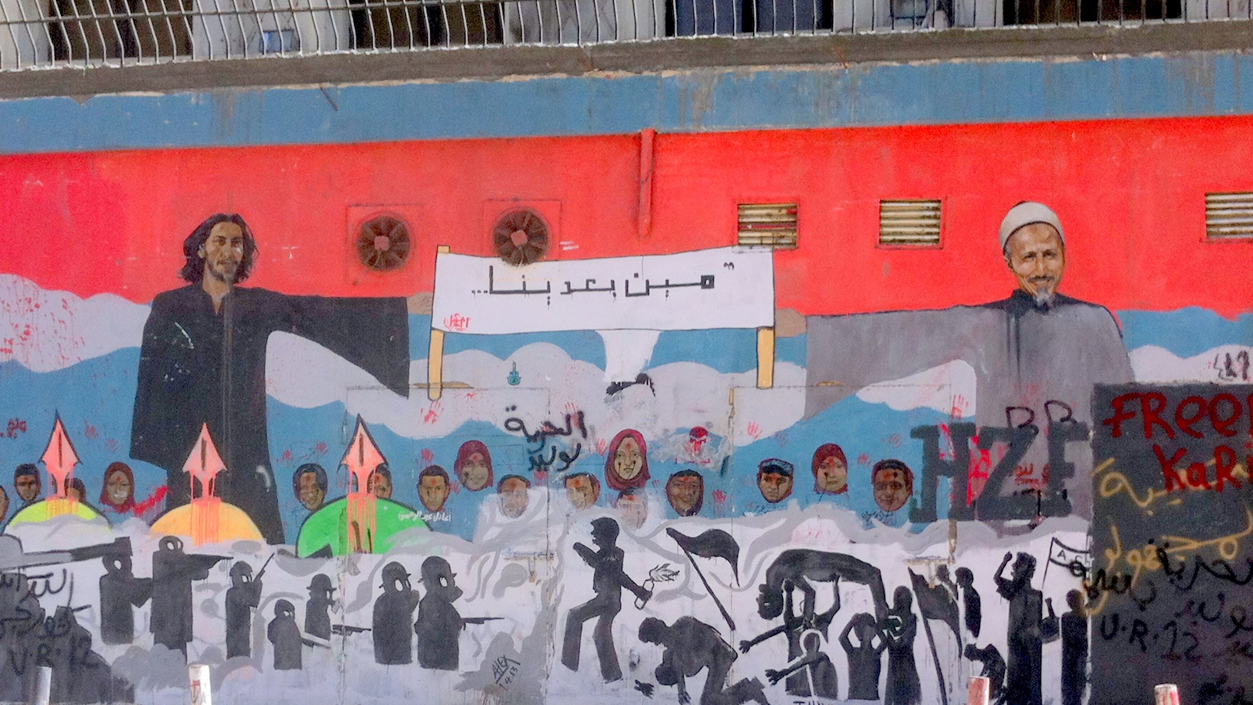 L'image montre une grande fresque murale qui illustre des figures humaines et des symboles de résistance. À gauche, on voit une personne avec de longs cheveux, et à droite, un homme âgé portant un turban. Entre eux, il y a une inscription dans une langue arabe. Le mural est riche en détails, avec des silhouettes de personnes et des flèches, suggérant des thèmes de lutte et d'espoir. Le fond est principalement rouge, créant un contraste fort avec les personnages et les éléments représentés.