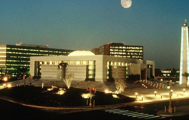 L'image montre un paysage urbain nocturne, avec un bâtiment moderne bien éclairé au centre. Ce bâtiment a une architecture distinctive, avec une coupole et des éléments de design contemporains. À l'arrière-plan, plusieurs autres structures se dressent, leurs fenêtres illuminées créant une ambiance vivante. On peut également voir une grande lune brillante dans le ciel, ajoutant une touche romantique à la scène. Des arbres et des zones paysagées entourent le bâtiment, et des lampadaires éclairent les chemins et les espaces extérieurs, rendant l'ensemble accueillant et esthétique.
