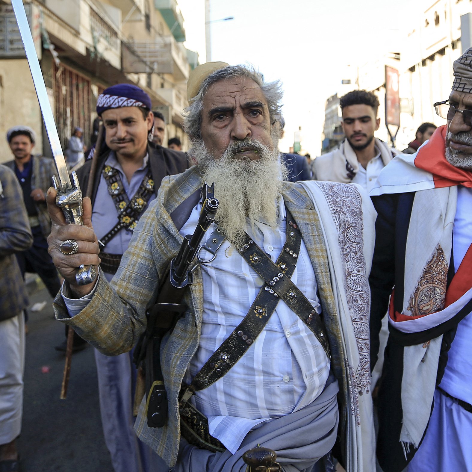 L'image montre un groupe d'hommes marchant dans une rue. L'un d'eux, au centre, est une personne âgée avec une longue barbe grise, tenant une épée. Il porte une tenue traditionnelle composée d'une chemise et d'une veste, ainsi qu'une ceinture. Les autres hommes dans le groupe portent aussi des vêtements traditionnels, et certains semblent porter des accessoires tels que des médaillons ou des fusils. L'ambiance de l'image suggère une manifestation ou une célébration culturelle. L'arrière-plan montre des bâtiments urbains typiques.