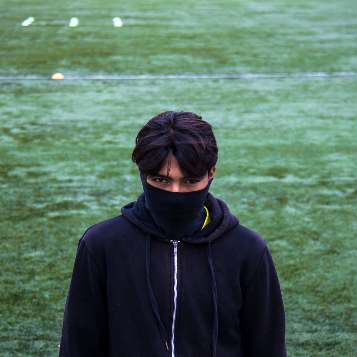 تظهر في الصورة شاب يقف على أرض ملعب رياضي مغطاة بالعشب الصناعي. يرتدي سترة سوداء ويغطي نصف وجهه بوشاح أو قناع أسود، مما يجعله يبدو غامضًا. نظرته مركزة، ويبدو جادًا في تعبيره، مما يضفي جوًا من القوة والتحدي على الصورة.
