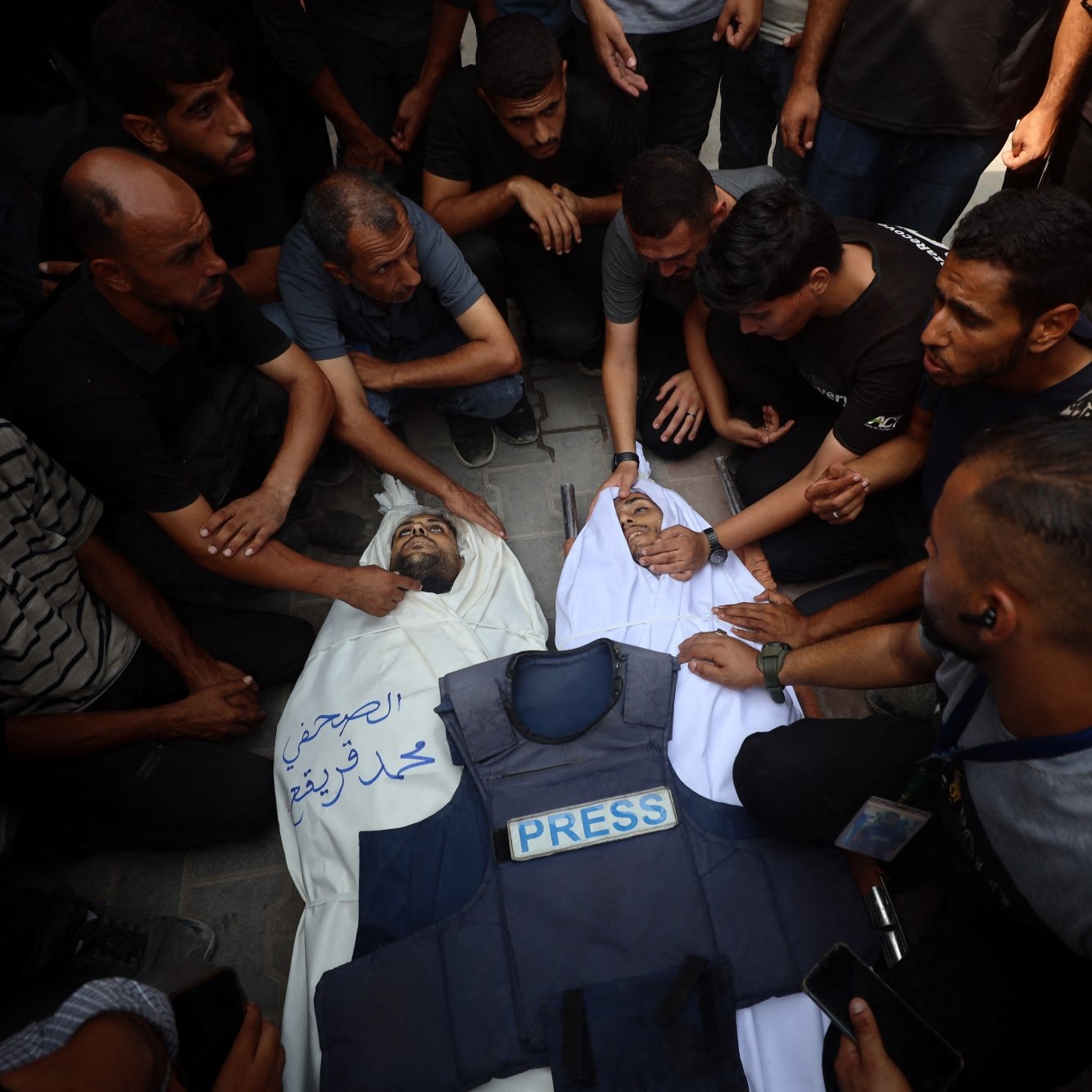A group of people mourns over a deceased individual in a white shroud, surrounded by a press vest.