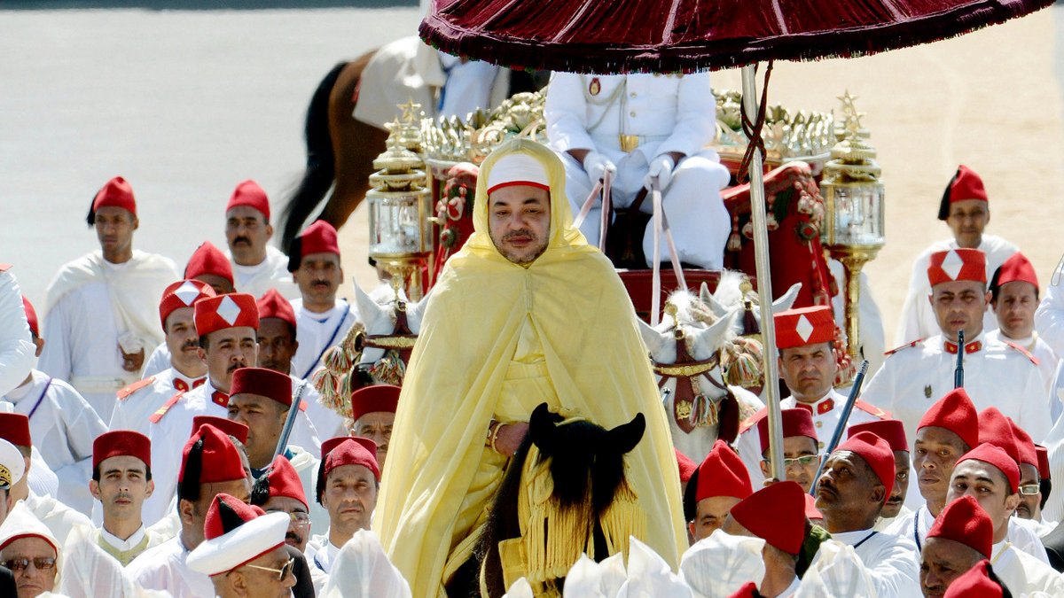 L'image montre un roi entouré de nombreux participants, tous vêtus de vêtements traditionnels. Le roi est monté sur un cheval, revêtu d'une cape jaune et portant un chapeau distinctive. Au-dessus de lui se trouve un parasol rouge. Les membres de l'entourage portent des turbans rouges, tandis que d'autres sont habillés en blanc. L'arrière-plan semble être une cérémonie officielle, avec des spectateurs et un décor sobre. Cette scène évoque une ambiance de tradition et de solennité.