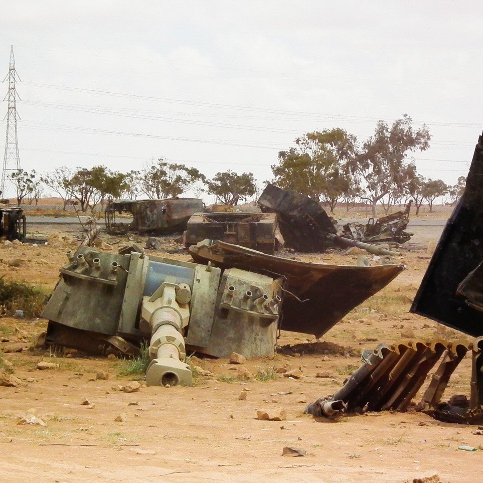 L'image montre un paysage désertique où sont dispersés des véhicules militaires rouillés et abandonnés. On peut voir des carcasses de tanks et d'autres machines, gisant sur le sol poussiéreux. Au loin, des arbres clairsemés et des pylônes électriques se distinguent, ajoutant à l'ambiance aride et désolée du lieu. Le ciel est nuageux, ce qui donne une atmosphère grise à la scène.