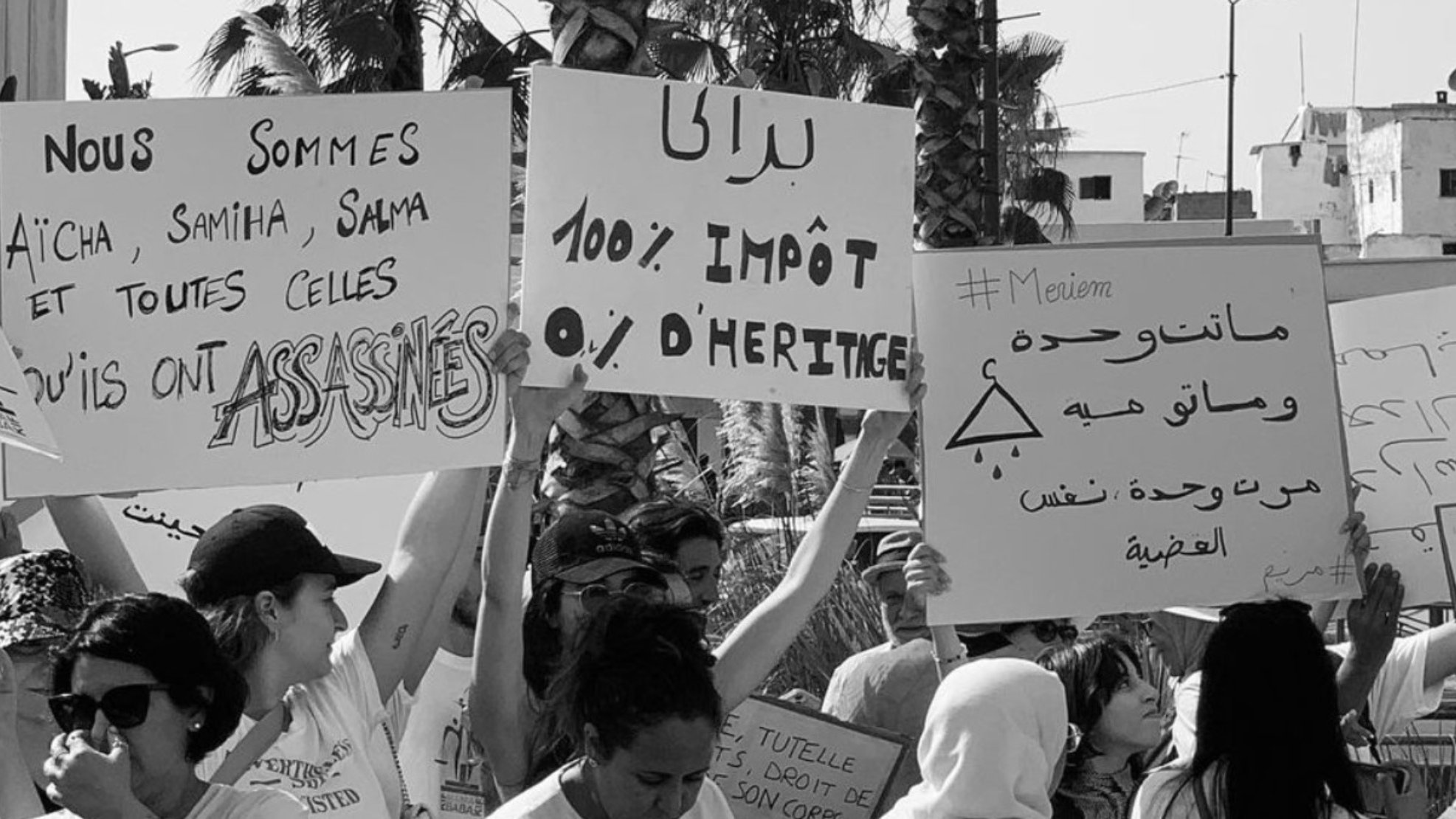 L'image est en noir et blanc et montre un rassemblement de personnes tenant des pancartes. Certaines des pancartes portent des messages en français comme : - "Nous sommes Aïcha, Samiha, Salma et toutes celles qu'ils ont ASSASSINÉES" - "100% IMPÔT 0% D'HÉRITAGE" Une autre pancarte comporte des inscriptions en arabe et le hashtag "#Meviem." Les personnes dans l'image semblent être engagées dans une manifestation ou une protestation concernant les droits des femmes et les questions de violence ou d'injustice. On peut également voir quelques palmiers et des bâtiments en arrière-plan, ce qui suggère que la scène se déroule en plein air dans un lieu urbain.