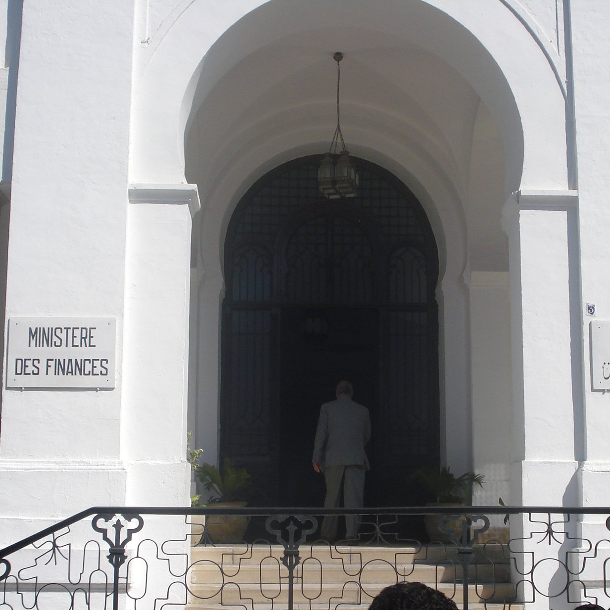 L'image montre l'entrée d'un bâtiment qui semble être le ministère des Finances, avec des inscriptions en français et en arabe. Le bâtiment est de style architectural classique, caractérisé par des murs blancs et des éléments décoratifs. On peut voir une grande porte d'entrée, entourée de fenêtres avec des grilles artistiques. À l'avant, une personne est visible, tournant le dos, s'approchant de l'entrée. Des plantes ornent l'espace autour des marches qui mènent à la porte.