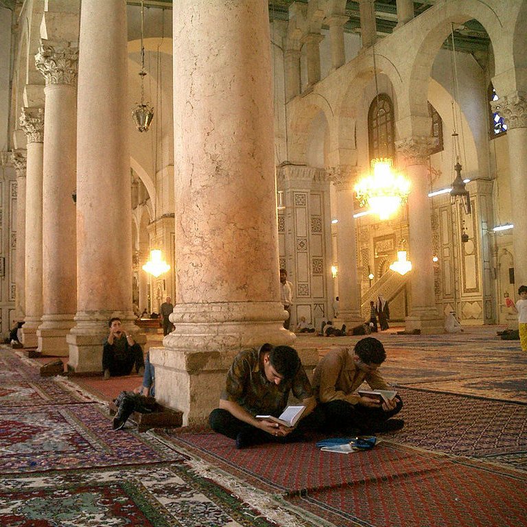 L'image montre l'intérieur d'une grande mosquée, caractérisée par de hauts plafonds et de majestueuses colonnes. Le sol est recouvert de tapis, et plusieurs personnes sont assises sur le sol, certaines concentrées sur leurs ordinateurs portables, tandis que d'autres semblent prier ou méditer. La lumière naturelle entre par les fenêtres, créant une ambiance calme et sereine. Des lustres pendent du plafond, ajoutant une touche d'élégance à cet espace sacré.