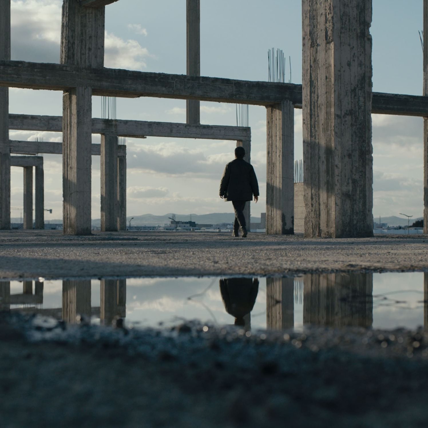 L'image montre une personne marchant à travers une structure de bâtiment inachevée. Les piliers en béton se dressent autour d'elle, créant une atmosphère à la fois vide et mystérieuse. Le sol est humide, et une flaque d'eau reflète les piliers et le ciel, ajoutant une dimension visuelle intéressante à la scène. Le ciel est partiellement nuageux, et l'éclairage est doux, contribuant à l'ambiance contemplative de cet espace abandonné.