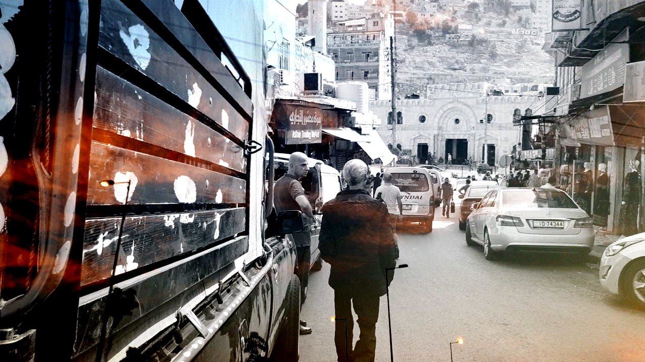 تبدو الصورة عبارة عن مشهد حضري يظهر شارعًا مزدحمًا حيث يسير أحد الأشخاص بعصا مساعدة. تظهر في الخلفية مباني متعددة الأشكال، وبعض السيارات تسير في الشارع. الأجواء تعكس حركة الحياة اليومية، ويبدو اللون الرمادي والأسود مسيطرًا مع لمسات من الألوان الدافئة، مما يضفي شعورًا بالحنين أو الذكريات.