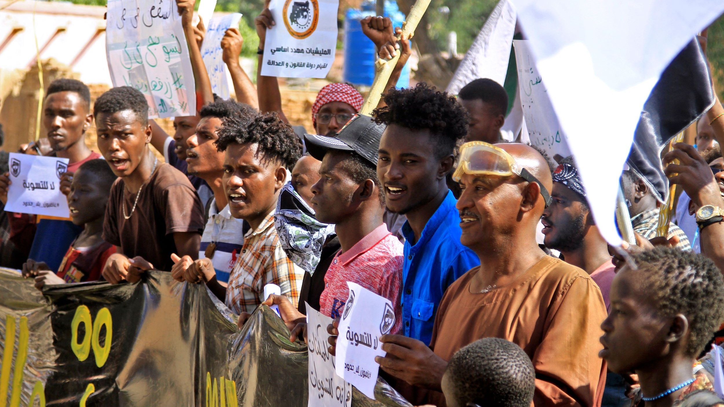 L'image montre un groupe de manifestants rassemblés, brandissant des pancartes et exprimant des slogans. Ils semblent engagés et passionnés, avec des expressions faciales intenses. Les gens portent des vêtements variés et il y a des drapeaux ou des bannières en arrière-plan. L'ambiance générale semble être celle d'une manifestation pour une cause précise.