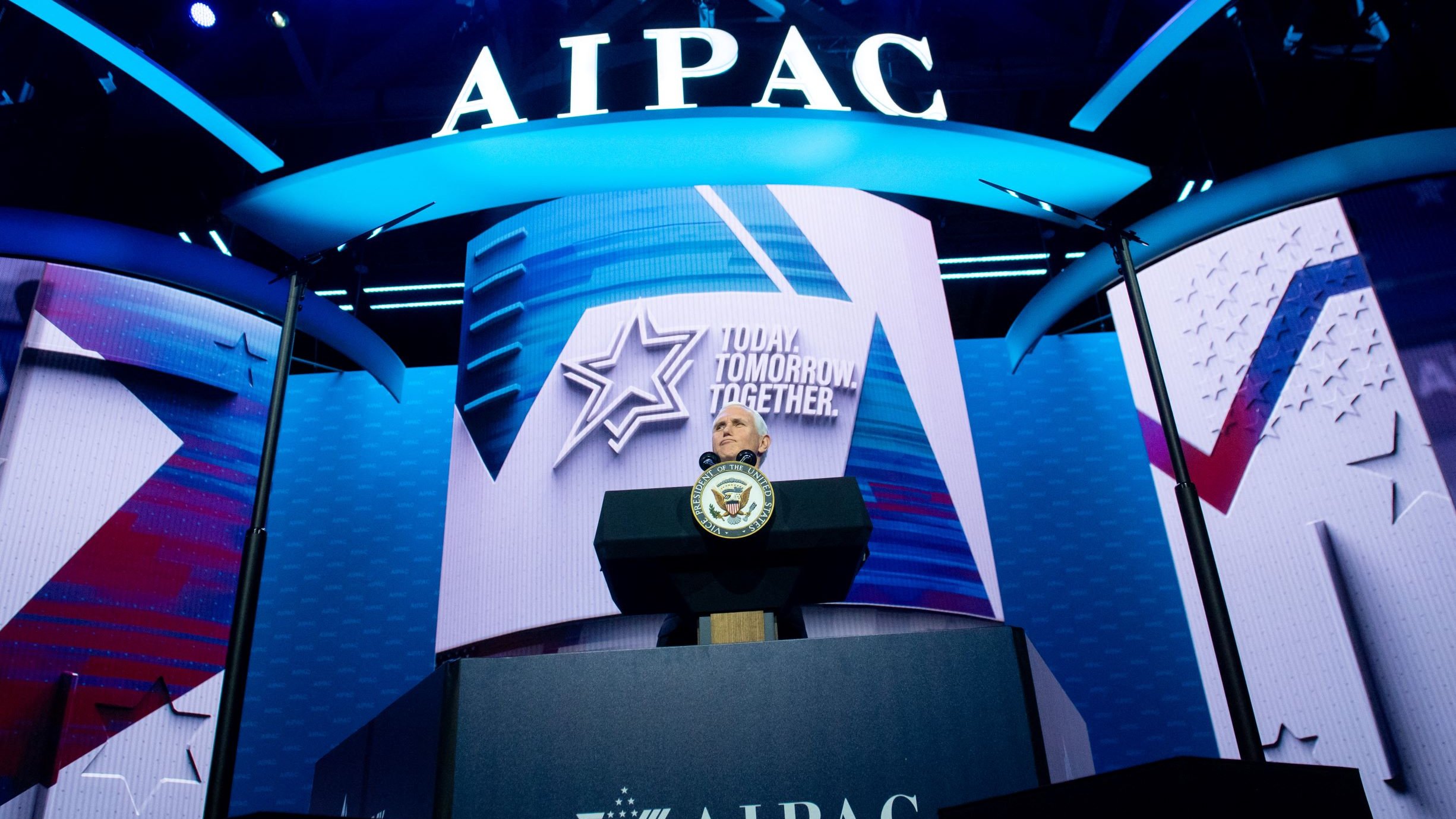 L'image montre une scène de conférence avec un podium bien en vue, sur lequel se trouve un sceau officiel. En arrière-plan, on peut voir un grand écran affichant le logo de l'AIPAC (American Israeli Public Affairs Committee), ainsi qu'un slogan qui pourrait être une thématique de l'événement. L'éclairage est dynamique, avec des lumières bleues qui créent une ambiance visuelle engageante. Cette configuration suggère qu'il s'agit d'un discours ou d'une présentation importante.