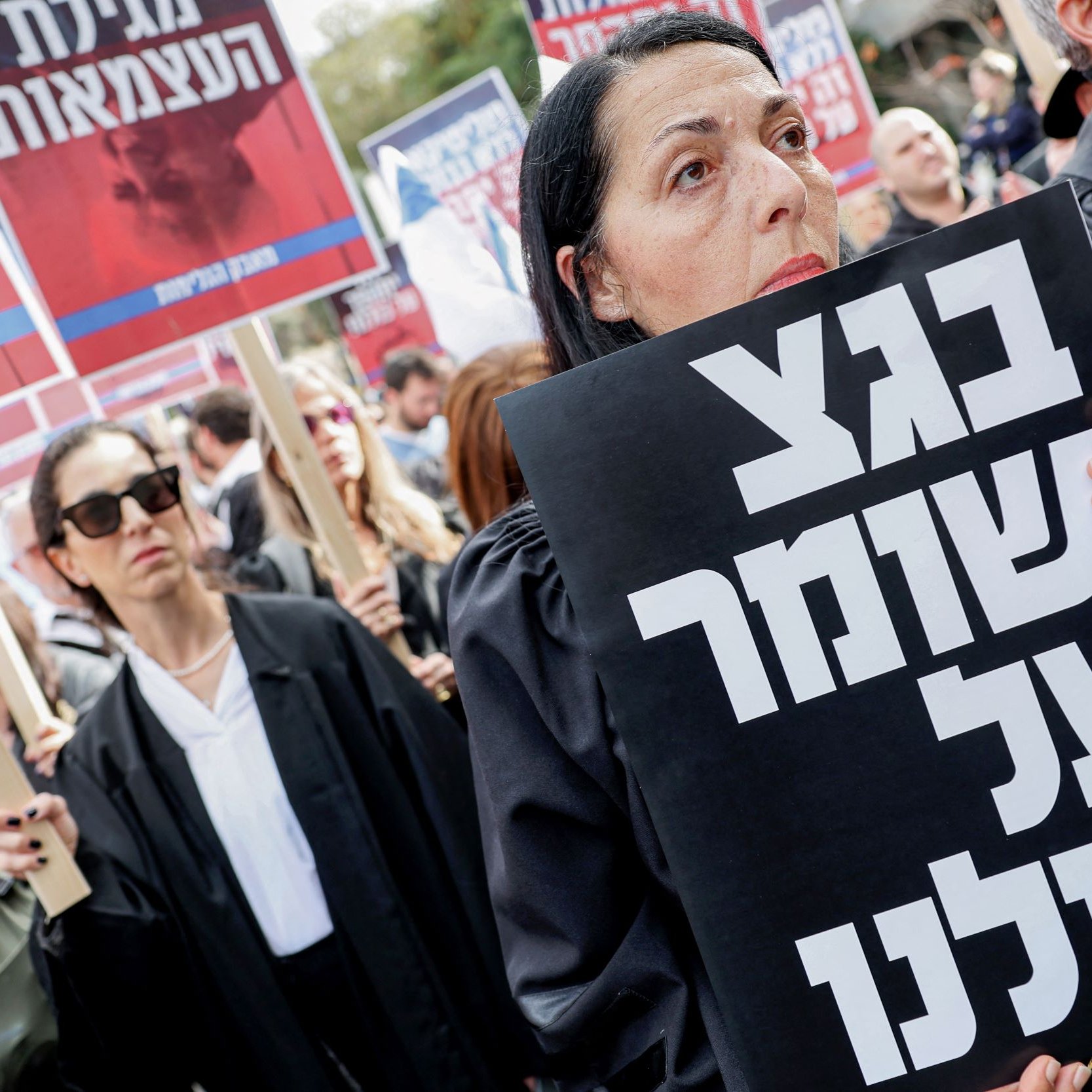 L'image montre une manifestation avec des participants portant des vêtements sombres, probablement des avocats, et tenant des pancartes. L'un des manifestants, au premier plan, tient une grande affiche noire avec du texte écrit en hébreu. L'atmosphère semble être sérieuse et engagée, avec des expressions résolues sur les visages des manifestants. En arrière-plan, d'autres personnes tiennent également des pancartes, ajoutant à l'ambiance de protestation.