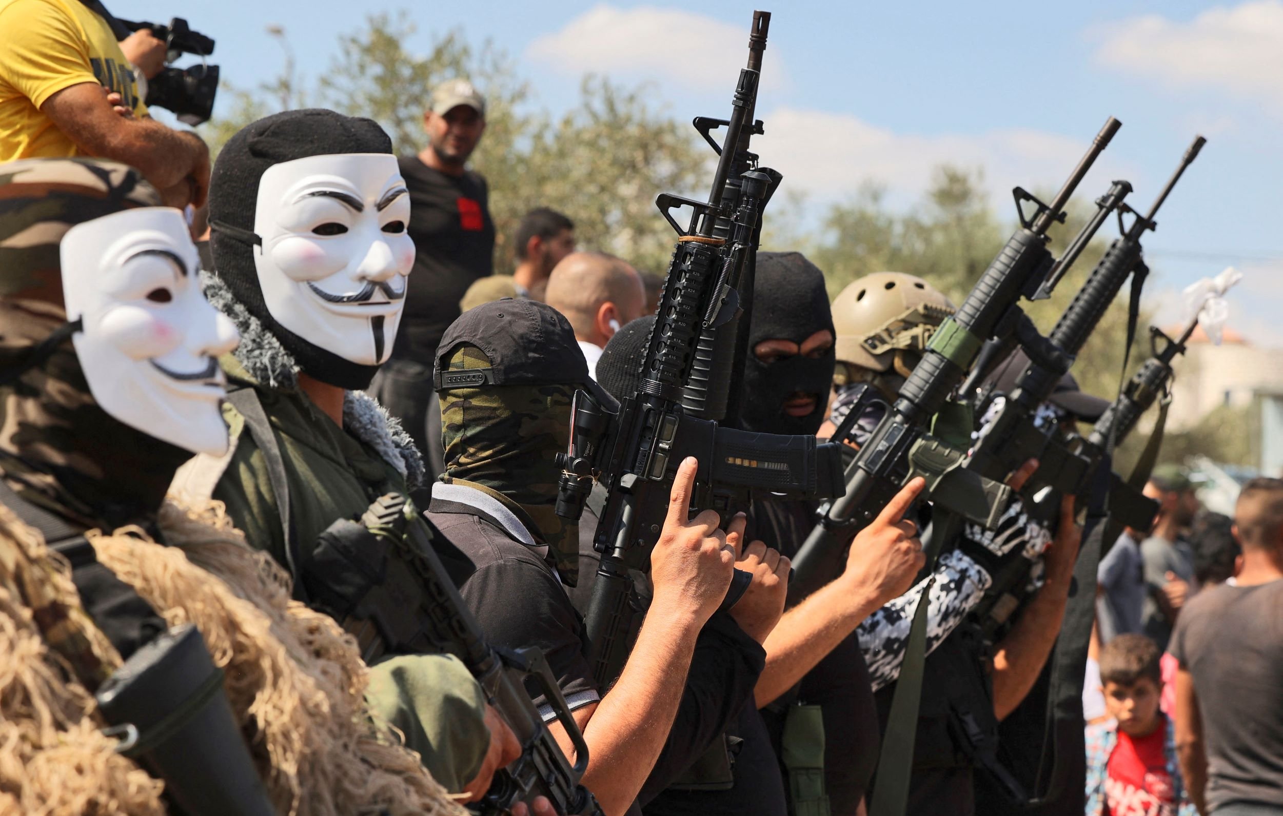 L'immagine mostra un gruppo di uomini che indossano maschere e abbigliamento militare. Sono schierati in formazione e tengono in mano diverse armi, alcune delle quali sembrano fucili. Sullo sfondo, si vedono altre persone che assistono alla scena. L'atmosfera sembra seria e intensa, e l'uso delle maschere suggerisce un intento di anonimato o di protesta.