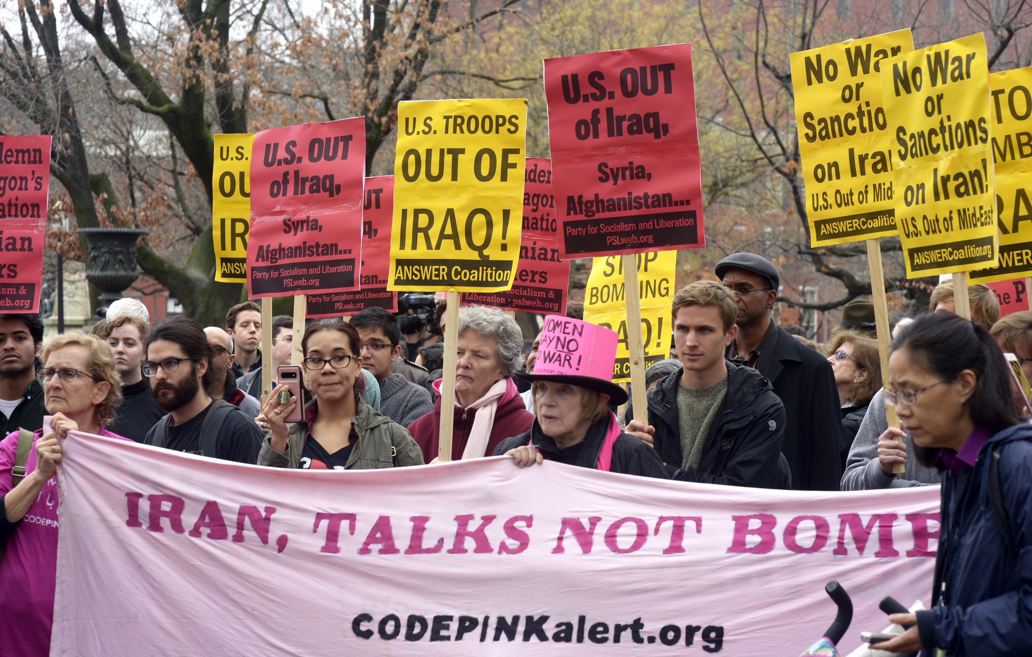 در این تصویر، گروهی از معترضان در حال برگزاری یک تظاهرات هستند. آنها پلاکاردهایی با نوشته‌های مختلف در دست دارند که بر ضد دخالت‌های نظامی ایالات متحده در عراق، سوریه و افغانستان و همچنین بر ضد تحریم‌ها و جنگ تأکید می‌کند. همچنین، در میان جمعیت، یک بنر بزرگ با نوشته «ایران، گفتگو نه بمب» مشاهده می‌شود. رنگ بیشتر پلاکاردها زرد است و جمعیت نشان از تنوع دارد. فضای تظاهرات جدی و متعهدانه به نظر می‌رسد.