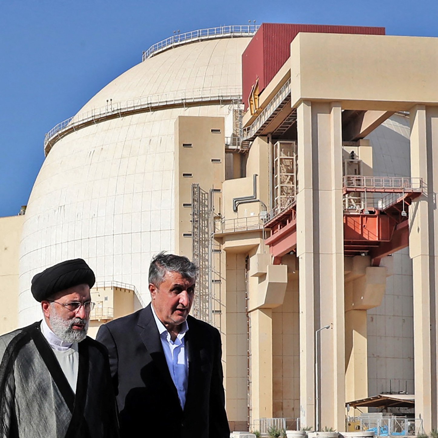 L'image montre deux hommes marchant devant une grande structure industrielle, qui semble être un réacteur nucléaire. Le bâtiment a une forme arrondie et est de couleur claire, avec des éléments de construction en béton visibles. Les deux hommes portent des vêtements formels ; l'un est vêtu d'un costume traditionnel et l'autre porte une veste. L'arrière-plan est dégagé, avec un ciel bleu. Cette scène pourrait illustrer une visite officielle ou une discussion liée à l'énergie nucléaire.