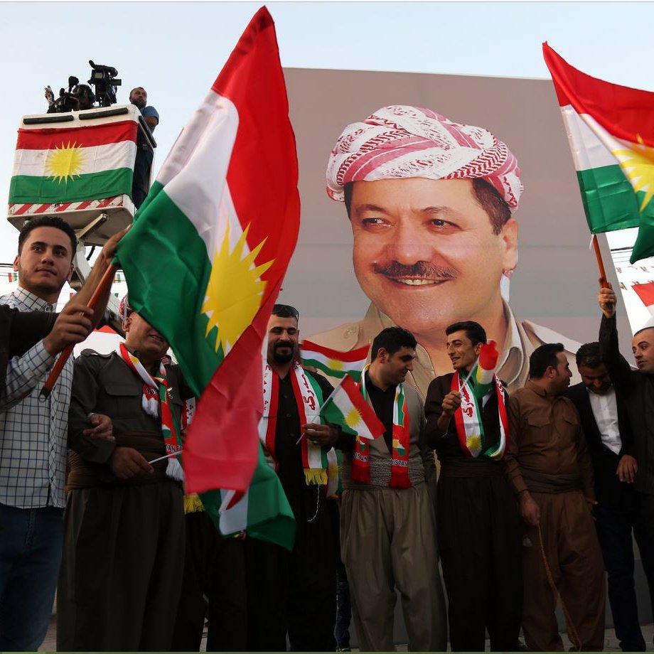 L'image montre un groupe de personnes tenant des drapeaux kurdes, célébrant devant une grande affiche d'une personnalité kurde. Les participants portent des vêtements traditionnels et semblent joyeux, suggérant une ambiance festive. L'affiche derrière eux représente un homme souriant, probablement un leader ou une figure importante dans la culture kurde. Les décorations et les drapeaux créent une atmosphère vibrante et patriotique.
