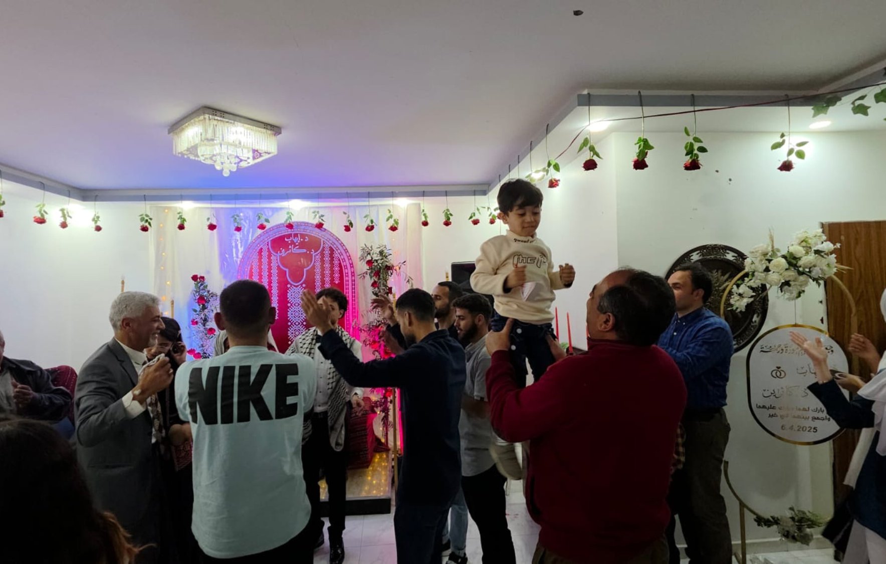 فضای شاد، مردم در حال رقص، تزئینات گل و نورپردازی رنگارنگ، یک کودک در آغوش بزرگترها.