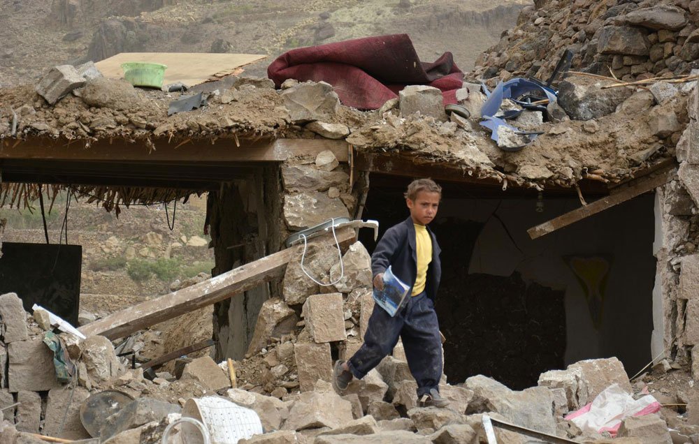 L'image montre un enfant marchant à travers des décombres d'un bâtiment détruit. Les murs sont effondrés et de nombreux débris, tels que des pierres et des objets éparpillés, sont visibles autour de lui. L'environnement semble être celui d'une zone touchée par un désastre, avec un paysage en arrière-plan qui pourrait suggérer des conditions difficiles. L'enfant porte des vêtements sombres, et son expression évoque une certaine gravité face à la situation environnante.