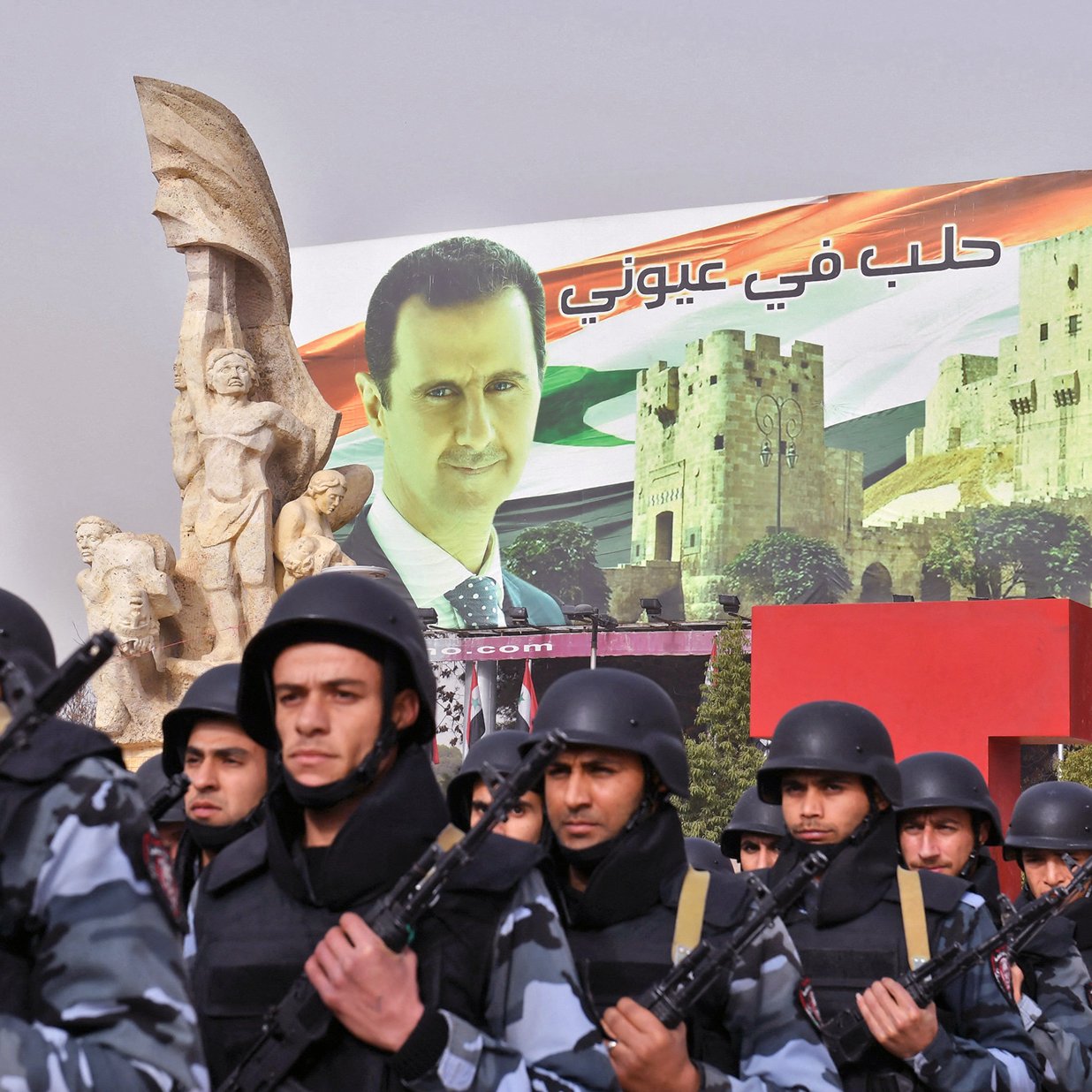 L'image montre un défilé militaire, avec des soldats en uniforme portant des casques et tenant des armes. En arrière-plan, on peut voir une grande affiche représentant un dirigeant, avec des bâtiments historiques et des éléments du drapeau syrien. La scène évoque une ambiance de démonstration de force et de soutien au gouvernement. Des sculptures monumentales sont également visibles, ajoutant une dimension artistique à l'ensemble.