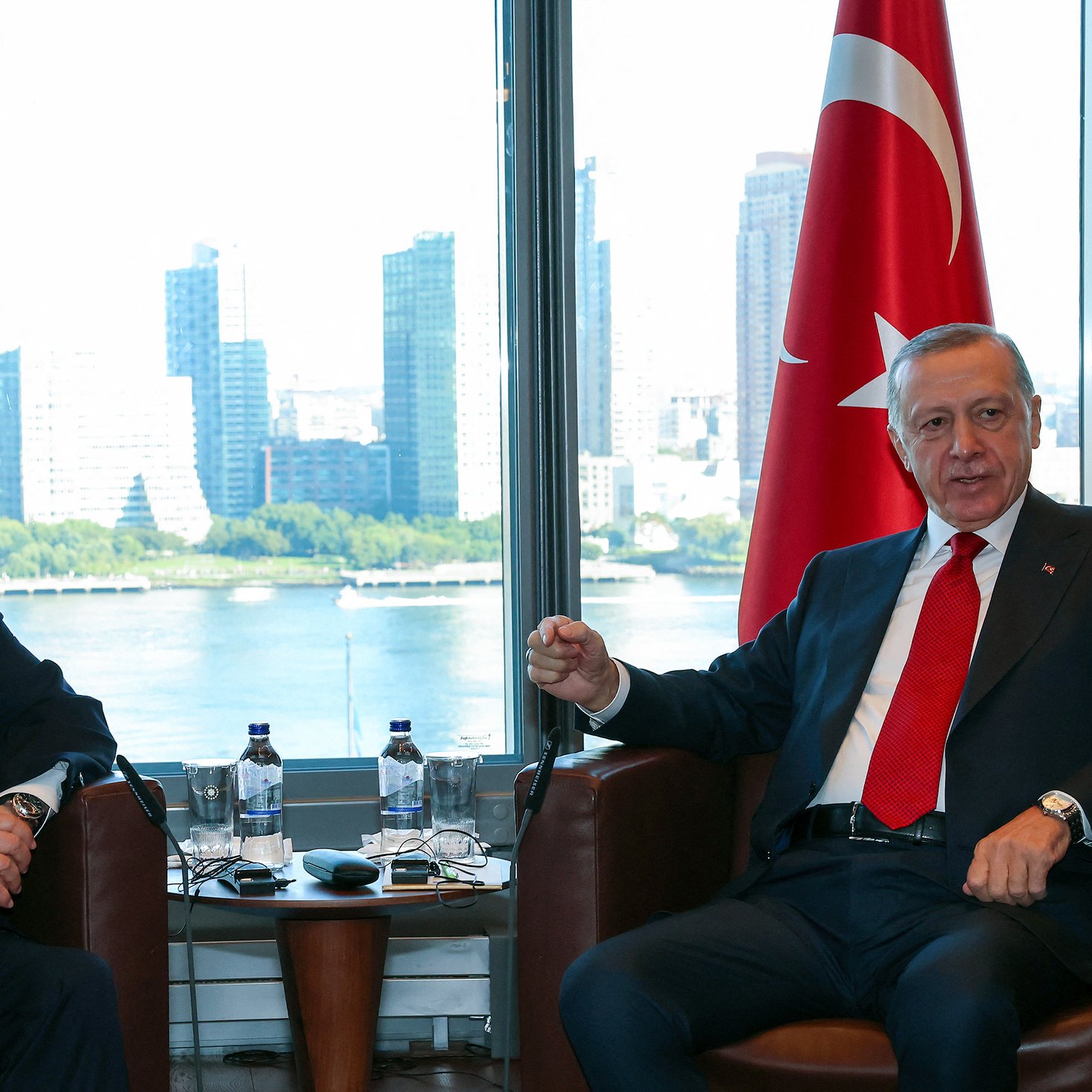 تظهر في الصورة اجتماع بين شخصين يجلسان في غرفة ذات إطلالة على مدينة حديثة. أحدهما يرتدي بدلة داكنة مع ربطة عنق حمراء، والآخر يرتدي بدلة رمادية. خلفهما يوجد علم إسرائيل إلى اليسار وعلم تركيا إلى اليمين. يظهر في الخلفية نهر وبعض المباني الشاهقة، مما يعكس أجواء حضرية حديثة. يبدو أن النقاش يجري في أجواء دبلوماسية.