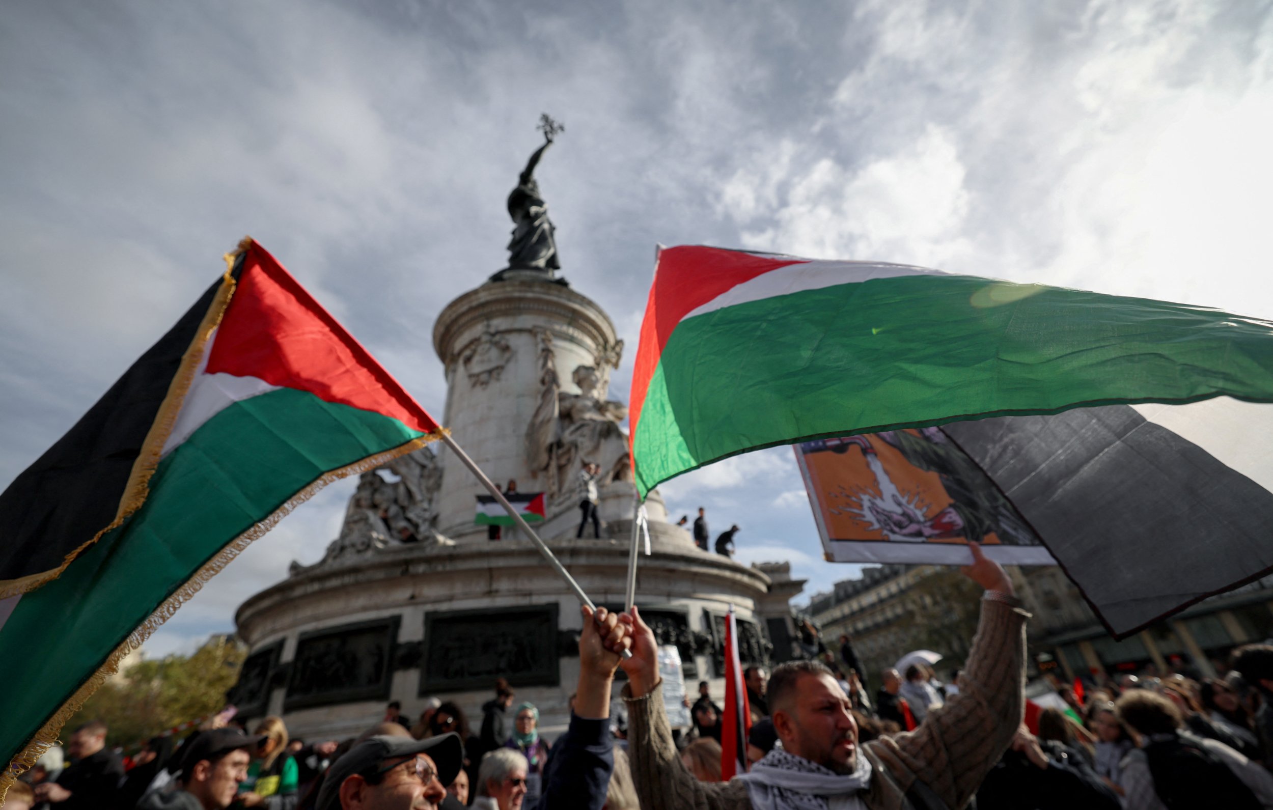 L'image montre une scène de manifestation avec des participants brandissant des drapeaux palestiniens. En arrière-plan, on peut voir un monument, probablement une statue, qui est entouré d'un ciel nuageux. Les personnes présentes semblent exprimant des sentiments forts, et l'atmosphère générale est celle d'un rassemblement public engagé et solennel.