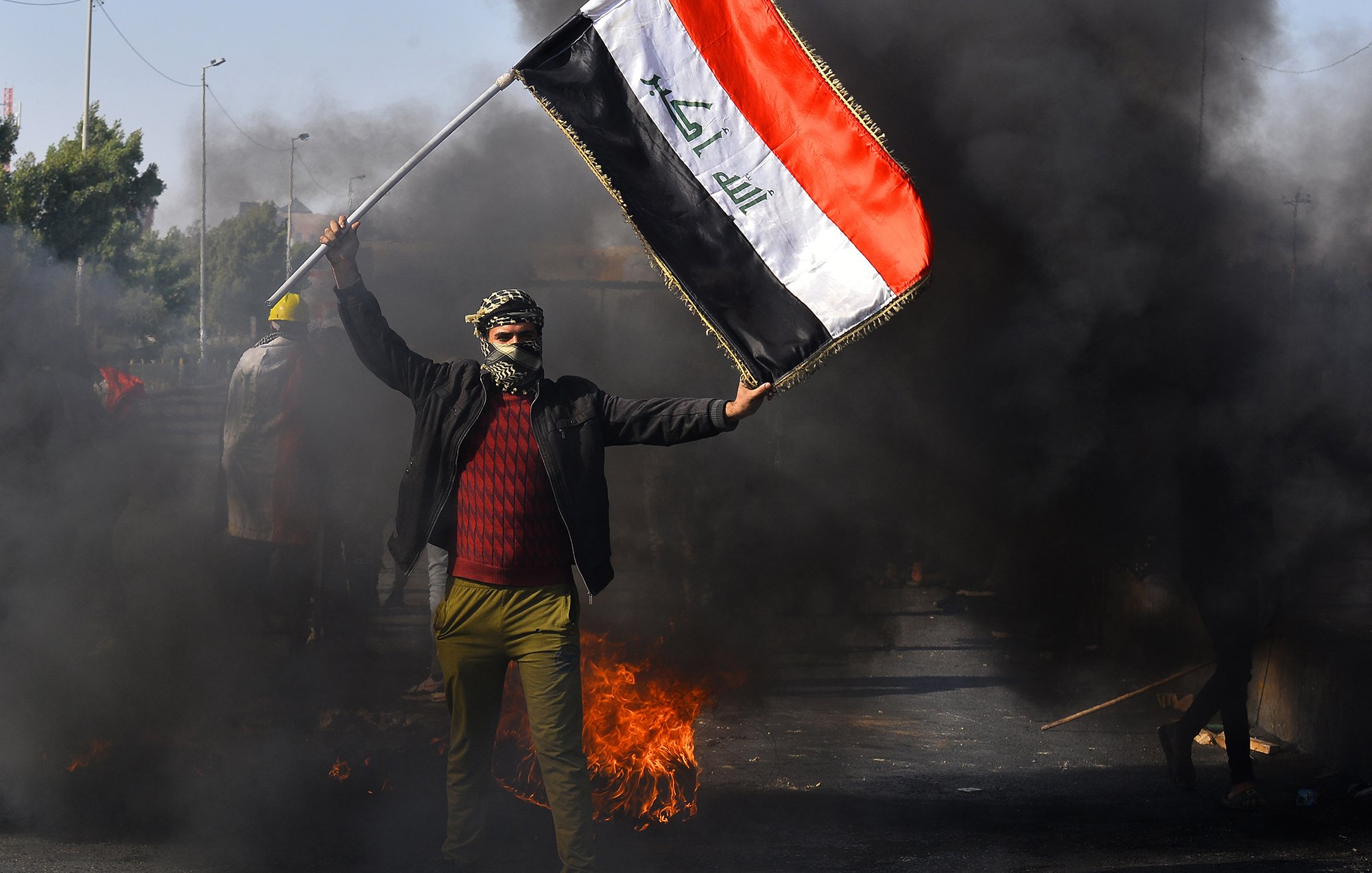 The image depicts a person holding the Iraqi flag amidst a scene of protest. The individual is wearing a mask and has their face partially covered, standing in front of billowing black smoke and flames. The background suggests a chaotic environment, likely indicative of unrest or demonstrations. The focus on the flag emphasizes national identity in the context of the protest.